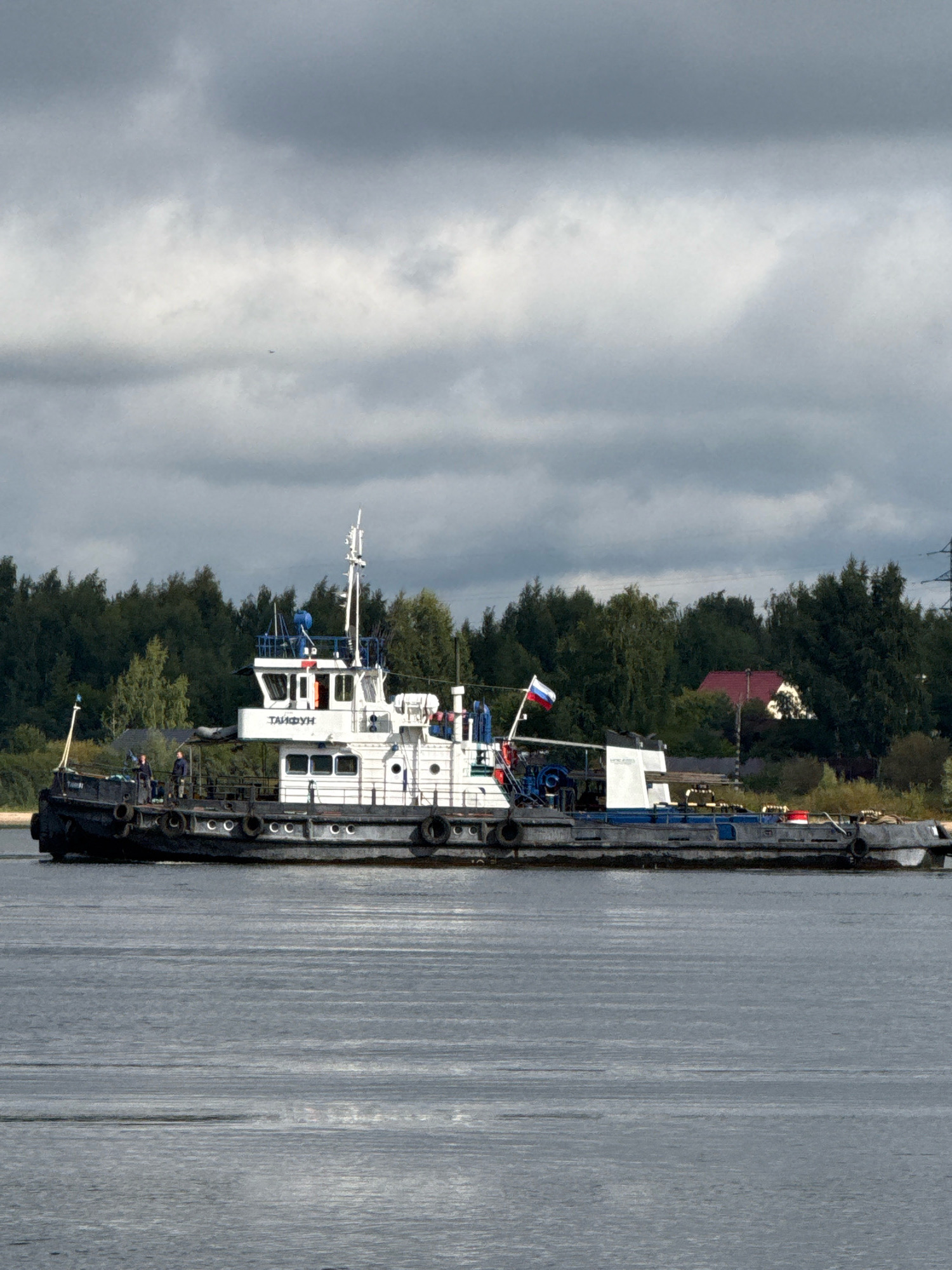A tugboat named "ТАЙФУН" is traveling on a body of water, likely a river, in Rusanovo, Russia. The boat is predominantly white and black with blue accents. Two individuals are visible on the deck of the tugboat. A Russian flag is flying from the vessel. The background consists of a treeline with a red-roofed building partially visible among the trees, and a power line tower in the distance. The sky is overcast with gray clouds. The water is calm with visible ripples.