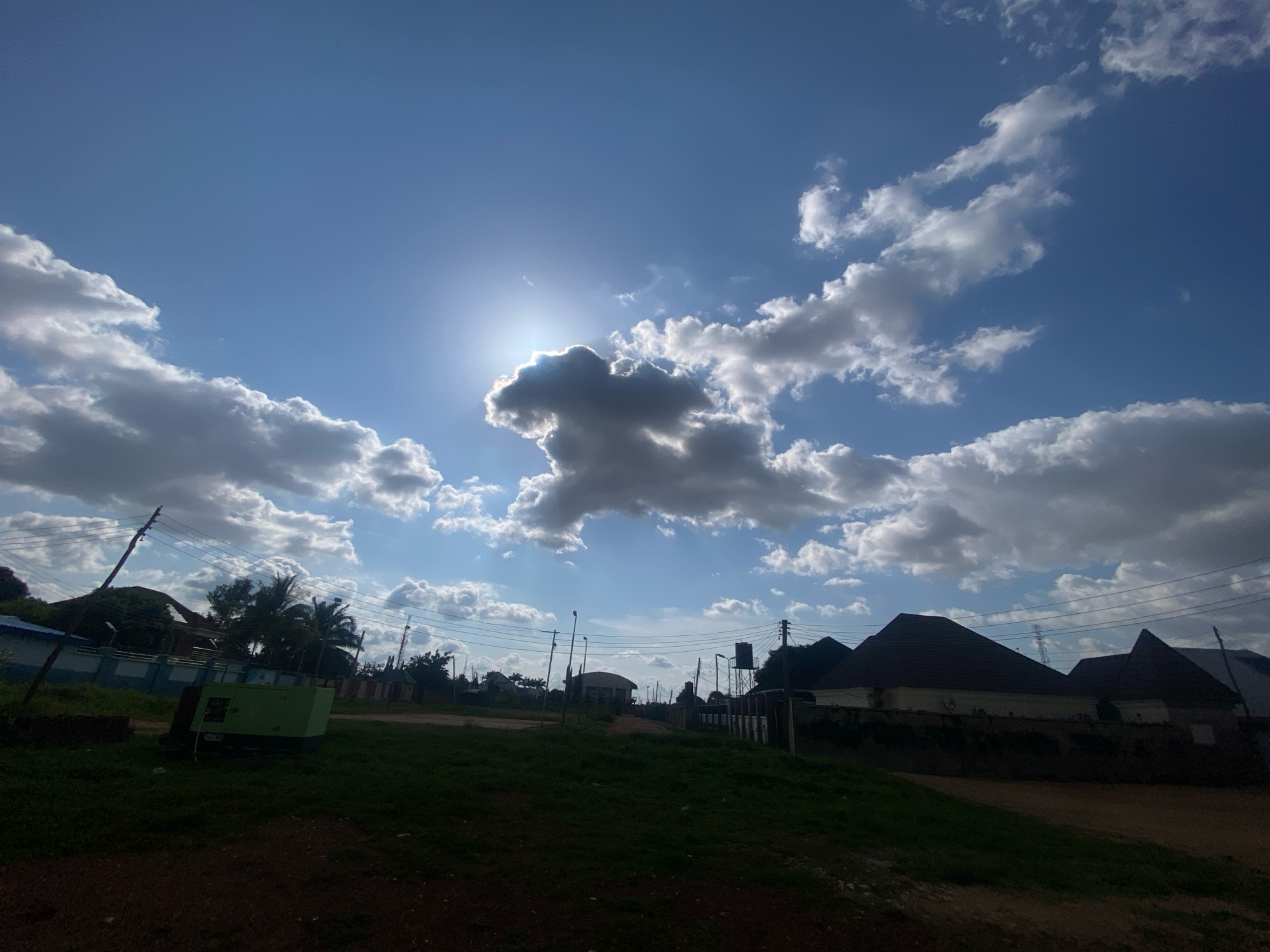 The image captures a street scene in Barden Yana, Nigeria, under a bright, partly cloudy sky. The sun is prominently visible, partially obscured by a large, dark cloud formation that creates a dramatic silhouette against the blue sky. Scattered white and gray clouds populate the upper portion of the frame.
In the foreground, a dirt path slopes upwards. To the left, a grassy area features a large, green generator. Beyond the generator, a wall with blue and white detailing runs horizontally, with a power pole and electrical wires extending upwards. A few palm trees and other vegetation are visible behind the wall.
Further into the scene, several houses with distinctive pitched roofs are situated along the street. These houses appear to be constructed with light-colored walls and dark-colored roofs. Electrical poles and wires are prevalent throughout the middle ground, indicating the presence of infrastructure. A water tank is visible atop one of the structures. The overall environment suggests a residential area within the city. No people are visible in the image.