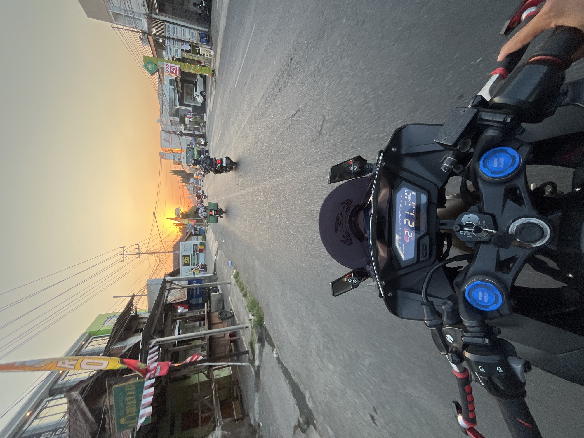 The image is a first-person perspective shot from a motorcycle on a street in Banyuputih, Indonesia, at sunset. The foreground shows the handlebars and dashboard of the motorcycle. The dashboard displays a digital readout of "221" and other unreadable data. The street is paved with asphalt and shows some wear and tear. On the left side of the street are buildings with shops and stalls, some of which are made of wood and corrugated metal. There are flags and signs visible on these buildings. On the right side of the street, there are more modern buildings, including one with a green "UGO 24" sign. Traffic is visible on the street, including several other motorcycles and a truck. The sky is a warm orange and yellow due to the setting sun. The overall scene depicts a typical street environment in Indonesia during the evening.