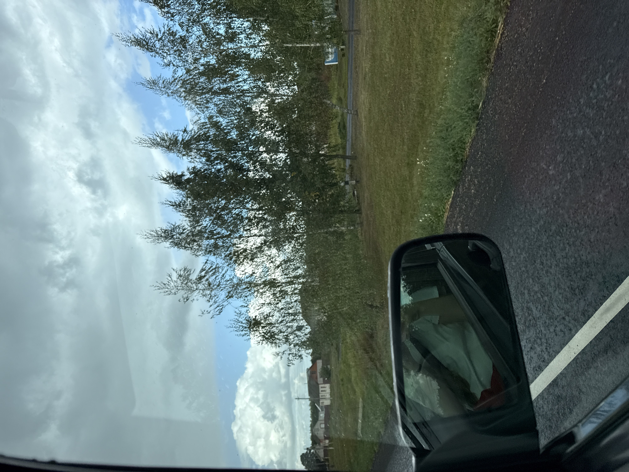 A view from inside a vehicle looking out the window shows a rural roadside scene. In the foreground, a side-view mirror reflects the interior of the car, showing a person wearing a light-colored shirt and red shorts. The road ahead is paved with asphalt and has a white line marking the lane. To the right of the road, a grassy verge transitions into a field with scattered trees. In the background, a line of birch trees with sparse foliage stands against a sky with a mix of clouds and blue patches. Further in the distance, there are several houses with red roofs, suggesting a village or small settlement. The overall impression is that of a travel scene in a moderately cloudy, daytime environment.
