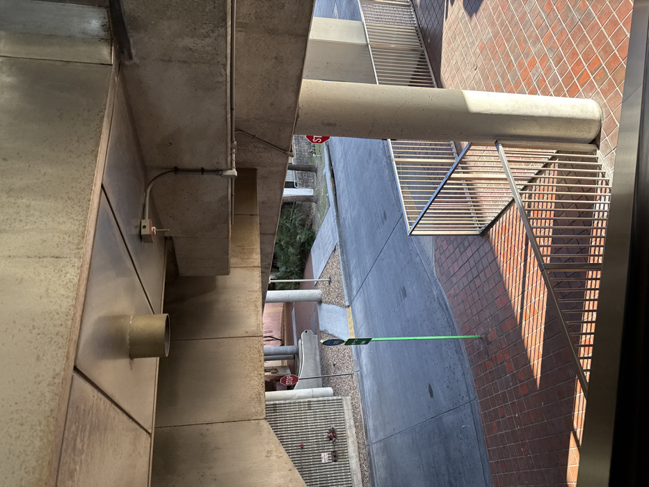 The image presents an elevated, downward-angled view of an outdoor public area located under an overhead concrete structure, identified as part of the Hialeah Market Station or Hialeah Station of the Miami-Dade Metrorail system in Hialeah, Florida, United States of America.
The foreground displays a reddish-brown square paver or brick surface, with strong parallel shadows cast across it by silver-colored metal railings that form a barrier or stairway descending out of frame to the lower left.
The mid-ground features a grey asphalt or concrete roadway. To the left of the roadway, a light grey concrete sidewalk transitions into a ramp. Adjacent to this, a vertical wall with a ribbed texture, possibly concrete or metal, features a red fire hydrant. A red hexagonal STOP sign on a dark pole is positioned near the left sidewalk. A light green pole, topped with a small dark blue and white sign, stands at the edge of the roadway. On the right side of the mid-ground, a second light grey concrete path or ramp is visible, bordered by sparse green vegetation including palm trees. A second red hexagonal STOP sign is partially visible behind a large, round concrete support column on the right.
The overhead structure consists of exposed, unpainted concrete beams and slabs. The underside of the ceiling includes metallic panels. A cylindrical, light-colored metal vent or pipe extends downwards from the ceiling. A red-tipped fire sprinkler head or sensor is mounted on a concrete beam, connected by a curved metal conduit. Additional metal conduits run along the concrete support beams. Multiple large, round concrete columns support the elevated structure, one prominently on the right side of the frame.
No individuals, vehicles, or animals are present within the depicted scene.