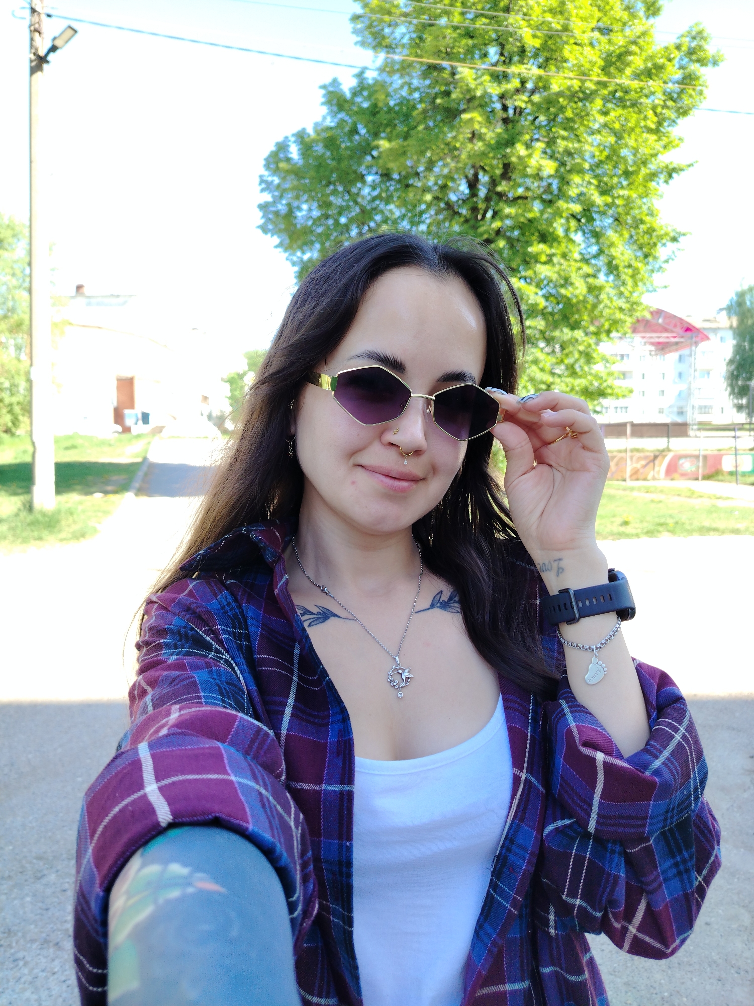 The image shows a person wearing sunglasses, a white tank top, and a plaid shirt. They have dark hair and visible tattoos on their chest and arm. The person is holding the sunglasses with their right hand. They are wearing a watch and a bracelet. In the background, there is a tree, a building, and a road. Based on the information available, the image appears to be taken in Noviy, Russia.