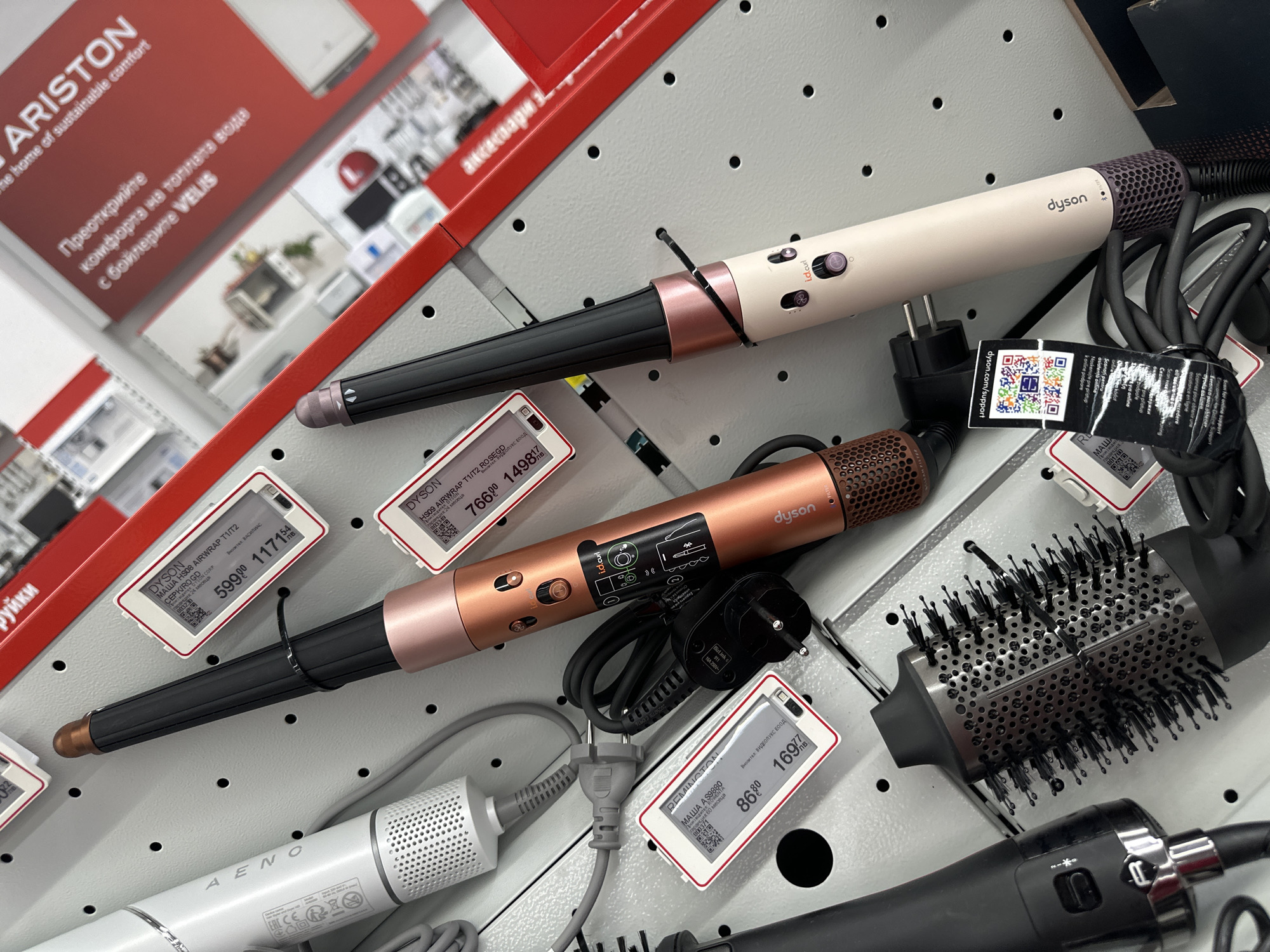 The image shows a retail display of hair styling tools, specifically Dyson Airwrap models and a Remington curling iron, arranged on a perforated metal panel in a store located in Lovech, Bulgaria. Price tags indicate the cost of the items in Bulgarian currency. A promotional poster for Ariston water heaters is visible in the background. The scene is brightly lit and seems to be inside a consumer electronics or department store.