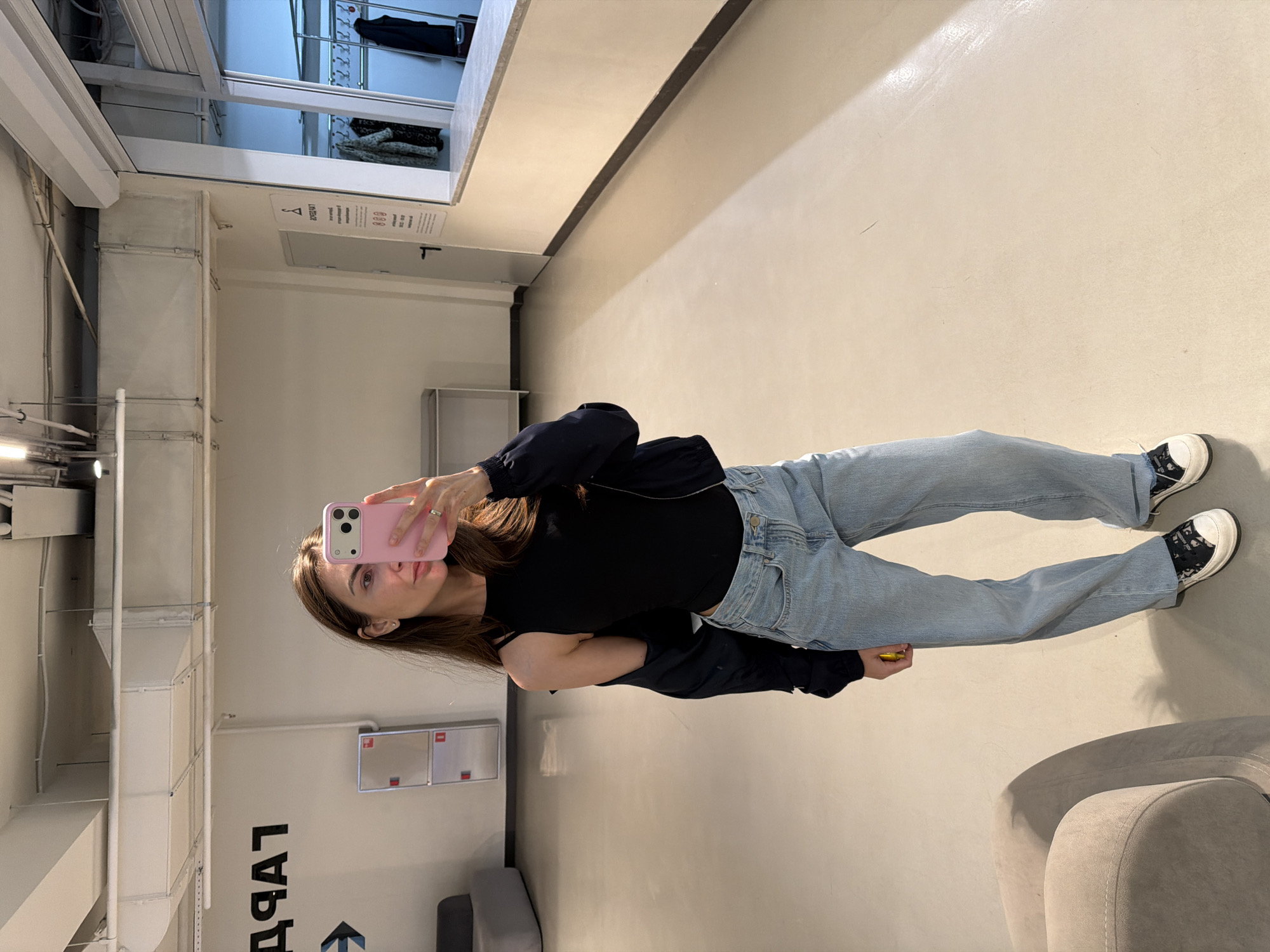 An image taken indoors shows a woman posing for a selfie in what appears to be a modern retail or public space. The woman, with long brown hair, is wearing a black long-sleeved top, light blue, loose-fitting jeans, and black and white sneakers. She is holding a pink smartphone up to her face with her right hand, capturing her reflection. A silver-colored ring is visible on her left hand's ring finger. She also appears to be holding a small yellow object in her left hand.
The setting features light-colored walls and a matching floor. Overhead, large industrial-style ventilation ducts and pipes are visible, extending across the ceiling and down a wall. In the background, shelves with folded clothes and clothing racks with hanging items can be seen, suggesting a clothing store or a fitting room area. A light grey upholstered armchair or sofa is partially visible in the foreground on the right.
Prominently displayed on a wall in large black letters is the Russian word "ГАРДЕРОБ" (GARDEROB), meaning "wardrobe" or "cloakroom," accompanied by an arrow pointing to the left. Another white information sign with a hanger symbol and some unreadable text is hanging from the ceiling. The overall lighting is artificial and bright. The woman maintains a neutral expression as she takes the photo.