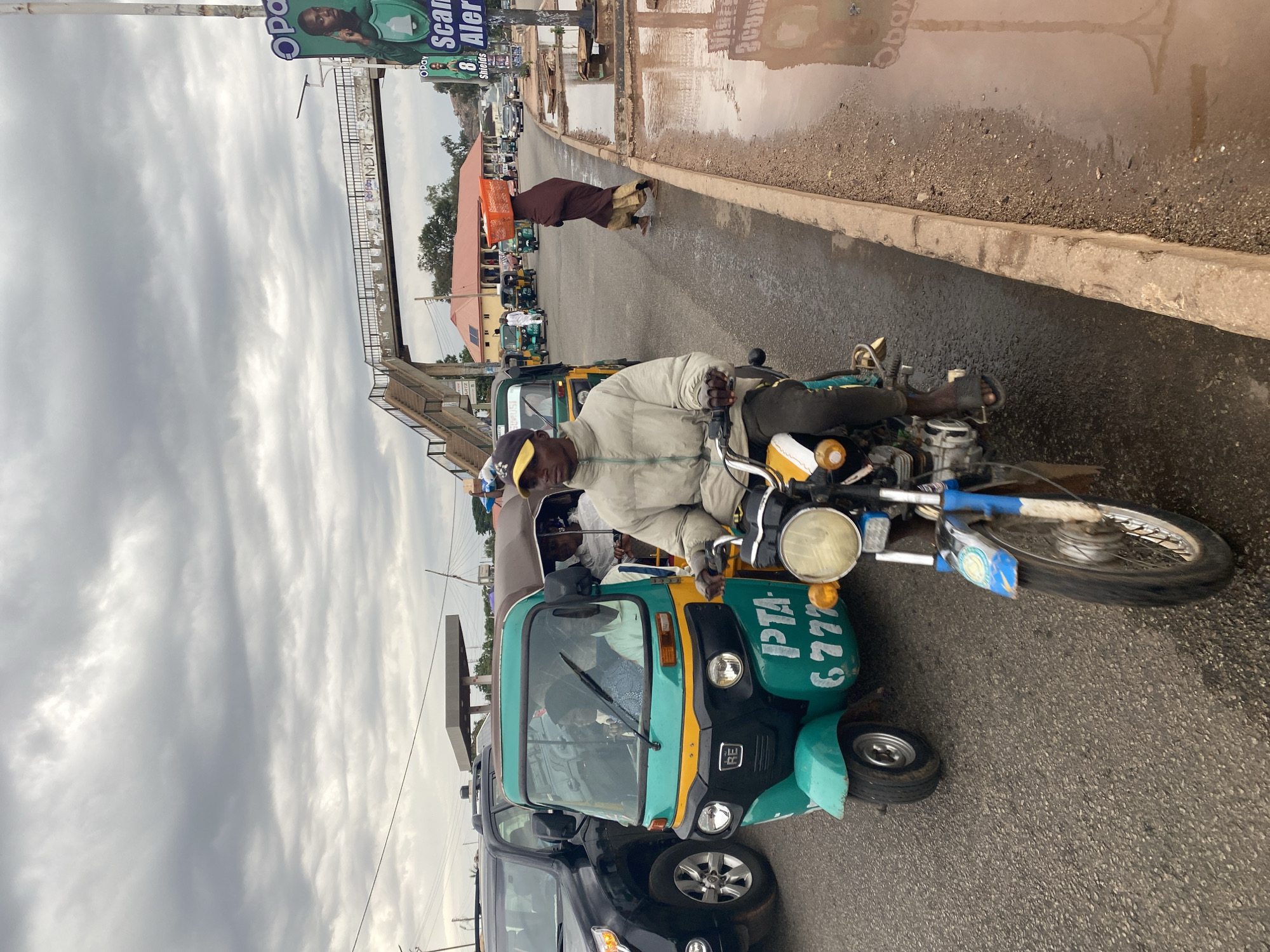 The image captures a scene in Gura Top, Nigeria. The foreground features a man riding a green and yellow auto-rickshaw (a type of motorized tricycle often used as a taxi). He wears a light-colored jacket and a cap. The auto-rickshaw has the writing "PTA 6777" on its side. A dark-colored car is parked behind the auto-rickshaw.
In the background, other auto-rickshaws, buildings, and people are visible on the street. There is a pedestrian bridge overhead, and a sign with the word "Opay" and the phrase "Scam Alert" can be seen on a pole. The street is wet, with puddles of water reflecting the sky. The sky is overcast with grey clouds.