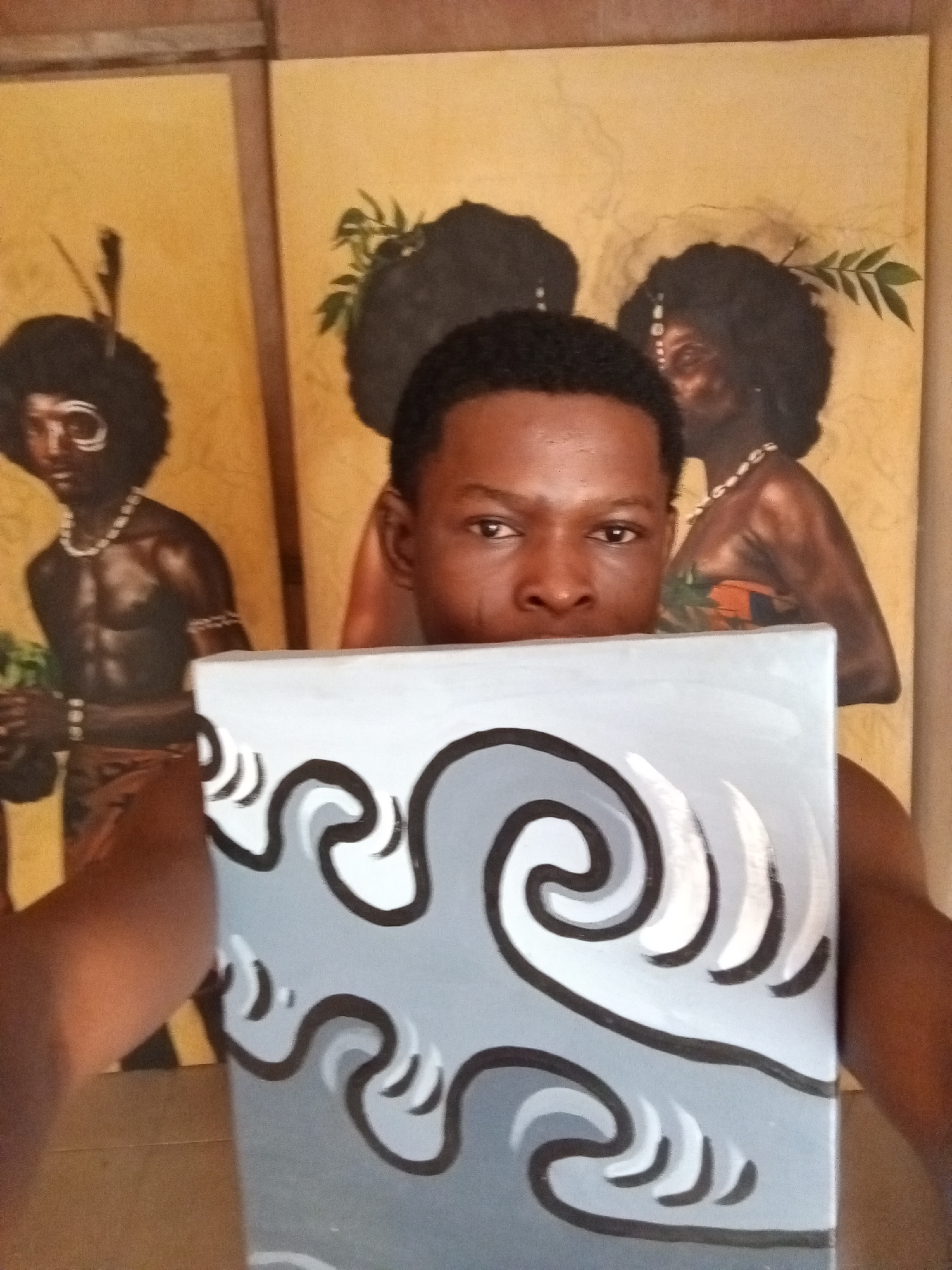 A medium shot captures a man indoors, holding a square canvas with a gray background and stylized, black-bordered wave patterns in shades of gray and white. The painting obscures the lower half of his face. Behind him are two large painted canvases. The left canvas depicts a bare-chested man with tribal markings and accessories. The right canvas shows two women with similar tribal aesthetics. The background color of both large canvases is a yellowish-tan. The location is likely an art studio or gallery in Ado, Nigeria.