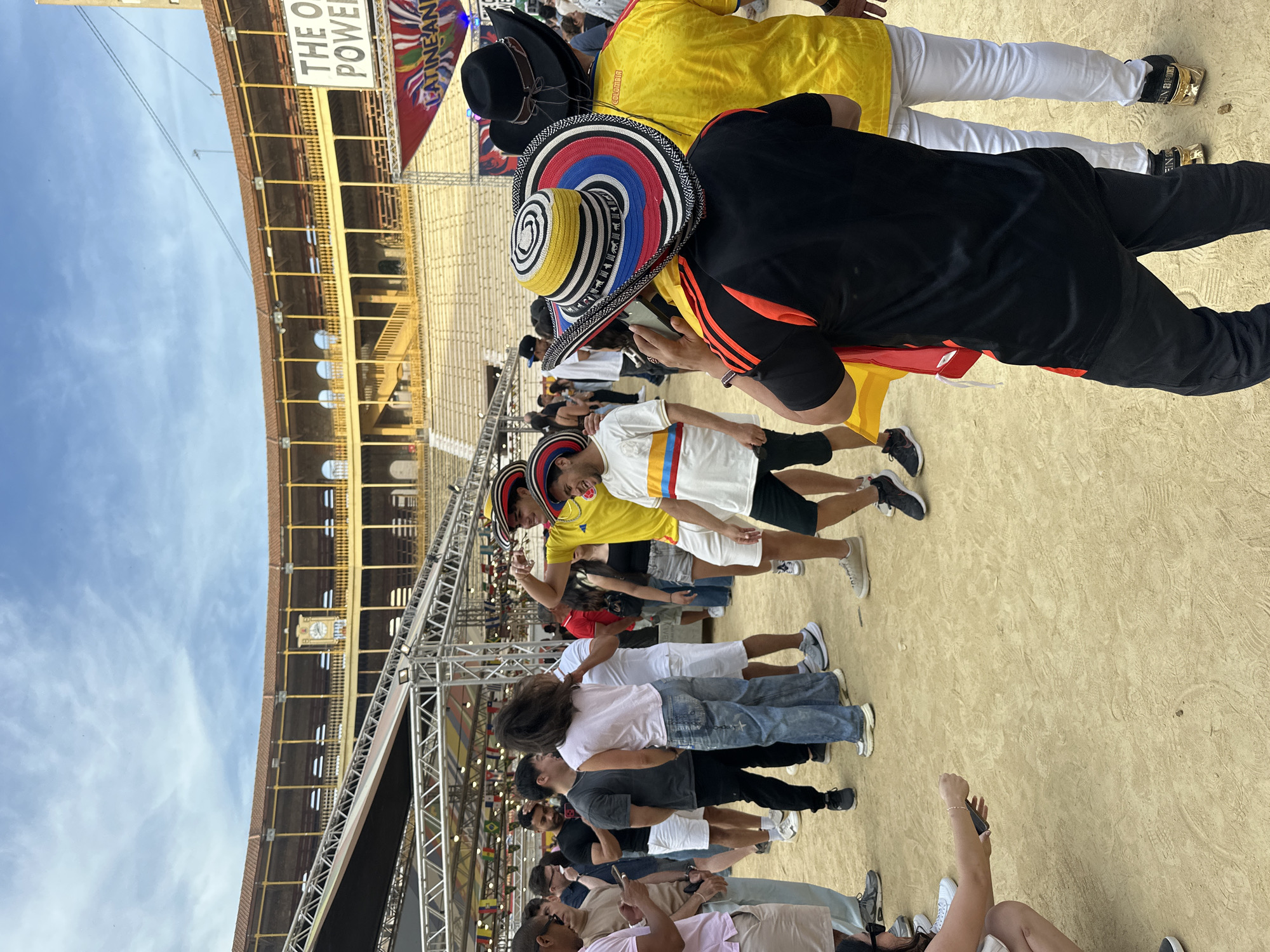This image captures an outdoor event within a bullring or arena, observed from the ground level. The background features the characteristic multi-tiered architecture of a stadium, with yellow balustrades, arcaded sections, and terracotta-colored roofs, all under a blue sky with scattered clouds. A large clock is visible on an upper tier of the stadium structure.
The foreground and midground are populated by numerous individuals gathered on the sandy, footprint-laden arena floor. Most attendees appear to be young to middle-aged adults dressed in casual clothing such as t-shirts, jeans, shorts, and sneakers. Several people wear distinctive hats: a person with their back to the camera, wearing a black t-shirt with orange details, sports a wide-brimmed sombrero vueltiao adorned with concentric circles of yellow, red, blue, black, and white stripes. Another individual in a yellow sports jersey wears a similar, predominantly yellow-striped sombrero vueltiao. A third person wears a black cowboy-style hat.
People are observed standing, conversing, and interacting with mobile phones. The individual in the black and orange shirt is holding a smartphone upwards, possibly recording. To the left, a woman crouches, holding a mobile phone. The event space is equipped with extensive metal scaffolding, supporting lighting rigs and banners, one of which partially displays "THE OLD POWER" and another "LATIN AMERICAN". Small flags are strung along the scaffolding. The stadium stands are visibly empty.