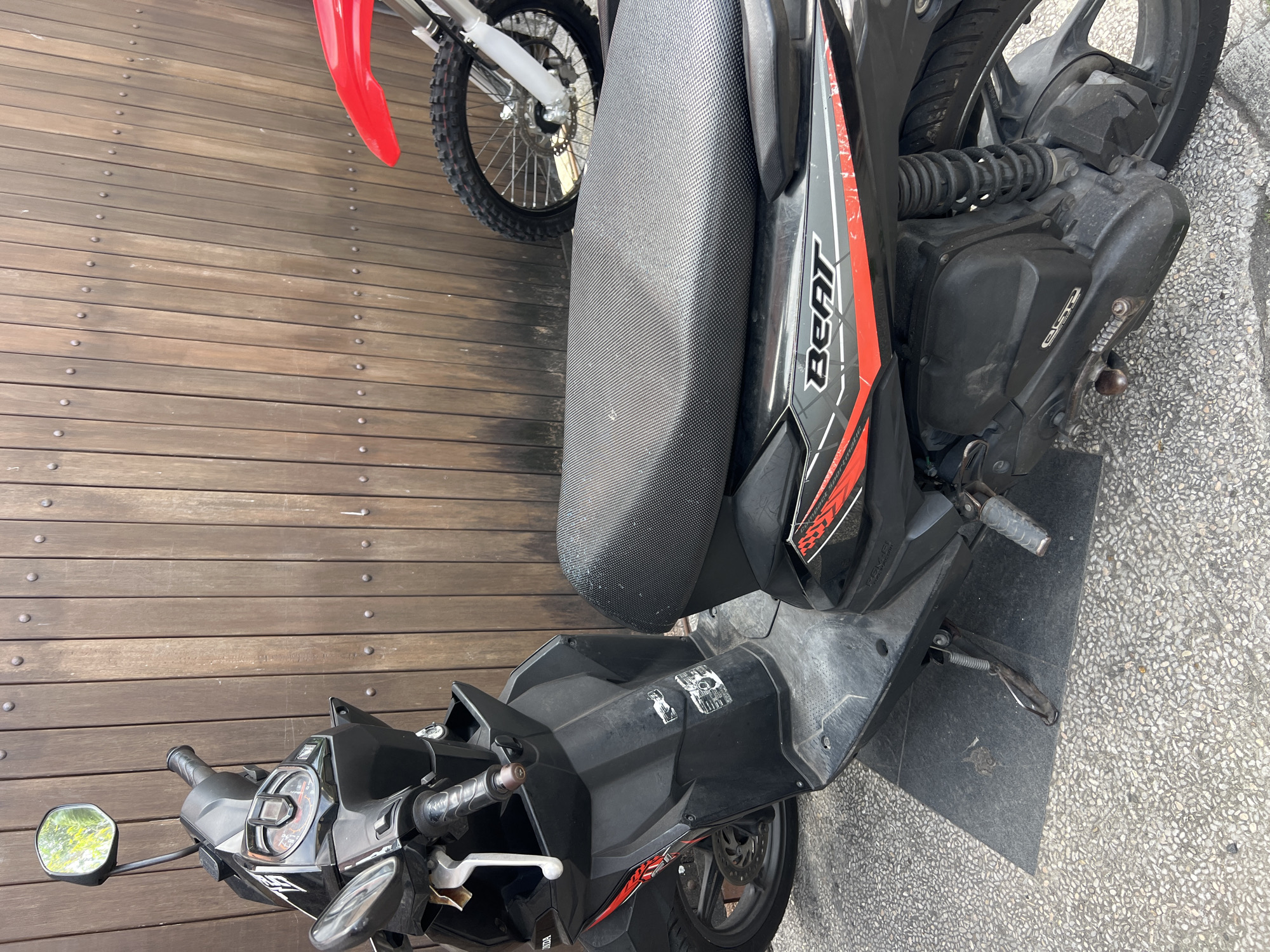A black Honda Beat scooter is parked on a paved surface in front of a wooden slatted fence. A red dirt bike's front wheel and fender are visible to the right of the scooter. The scooter has a textured black seat with a red and grey "BEAT" decal. There are some stickers on the scooter's body. The environment appears to be outdoors, possibly a street or alley in Denpasar, Indonesia, given the typical urban setting for such vehicles and architecture. No people are present in the image.