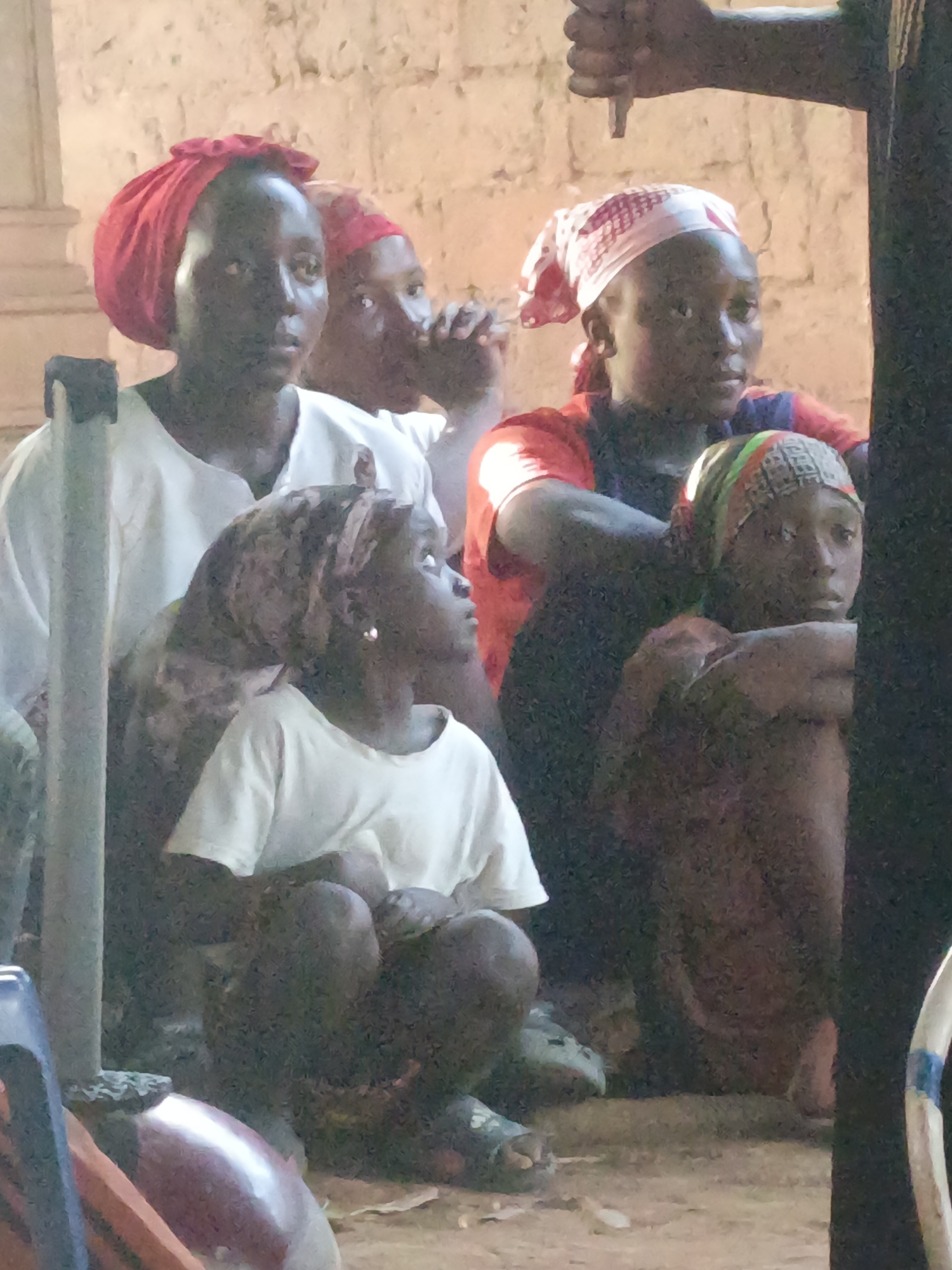 Akwanga, Nigeria: Five individuals are seated on the ground, attentively observing an event occurring outside the frame. The individuals are dressed in casual clothing, with red and patterned head coverings. A brick wall serves as a backdrop. A hand, possibly holding an object, is visible in the upper right corner. Furniture is scattered around.