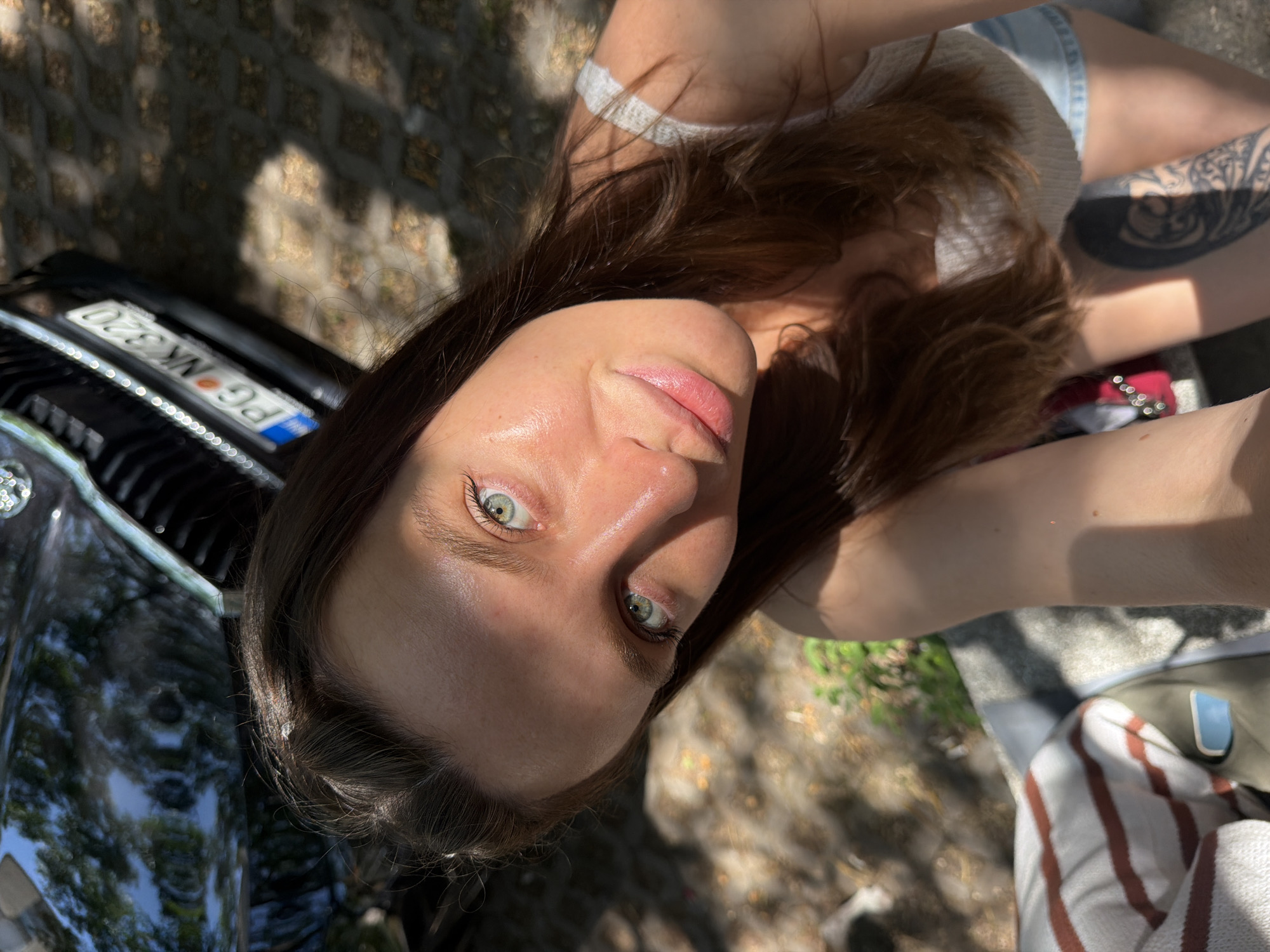 The image is a low-angle selfie of a young woman with light skin and brown hair. She has prominent green eyes and her face is in focus. She is wearing a light-colored tank top and denim shorts. A tattoo is visible on her upper right thigh.
In the background, there is a black car with a license plate that reads "PG NK 320" on it. The surrounding environment appears to be outdoors, with dappled sunlight and shadows cast by trees on the ground, which suggests an urban or park-like setting. Given that the question references Budva, Montenegro, it can be assumed that the image was taken somewhere in that city. However, with only the image to go on, the exact location cannot be determined.
Other objects visible include what appears to be a striped towel and a bag, likely situated on the ground to the right of the woman. The primary action is the woman taking a selfie.