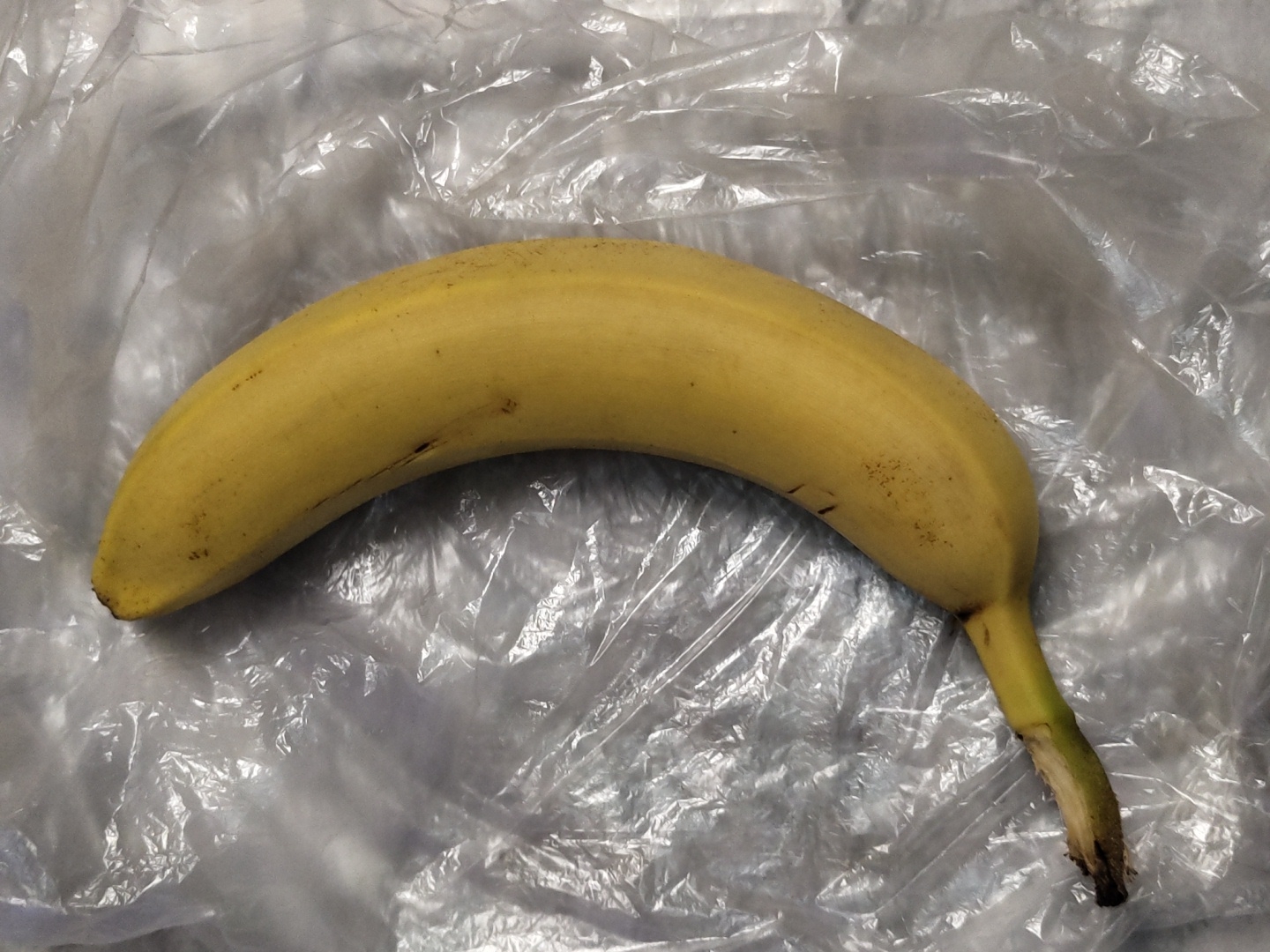 A single, ripe yellow banana is centrally positioned, curving from the bottom-left to the upper-right of the frame. Its skin exhibits a few small dark brown speckles and minor surface abrasions. The stem end, located at the bottom-left, is a pale greenish-yellow, culminating in a dried, dark brown/black tip. The distal end (top-right) also features a small, dark brown/black cap. The banana rests on a highly textured, translucent plastic sheet or bag, which is extensively crumpled and folded, creating irregular patterns of specular highlights and shadowed creases across a predominantly light gray and white surface. The lighting appears to be diffuse overhead, emphasizing the plastic's relief and the banana's form. No individuals or additional objects are present in the frame. The scene is located in the city of Unknown, Unknown.