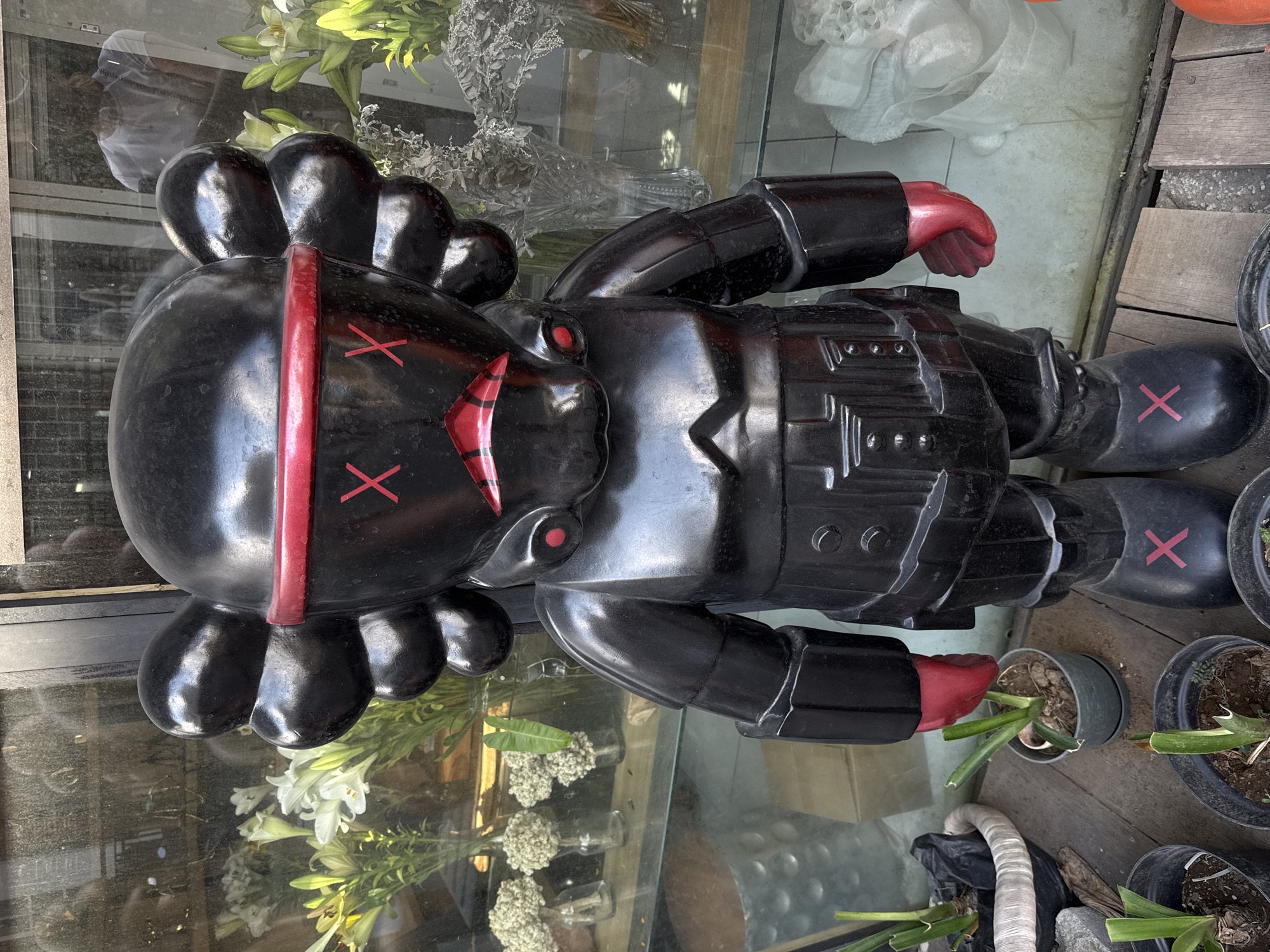 In the city of North Kuta, Indonesia, a large, black, stylized cartoonish figurine stands on a wooden surface. The figurine has a black helmet-like head with red detailing around the eyes and mouth. The eyes are marked with red "X" shapes, and the mouth is a red, downturned shape also resembling "X"s. Red gloves cover its hands. Its body is black, with metallic-looking details on the chest and arms, and red "X"s are visible on its black boots. Behind the figurine, through a glass pane, a display of white and yellow flowers in vases can be seen, along with other floral arrangements. The environment appears to be an outdoor setting, possibly a shop or a market stall, with more potted plants visible around the figurine's base. The lighting suggests it is daytime.