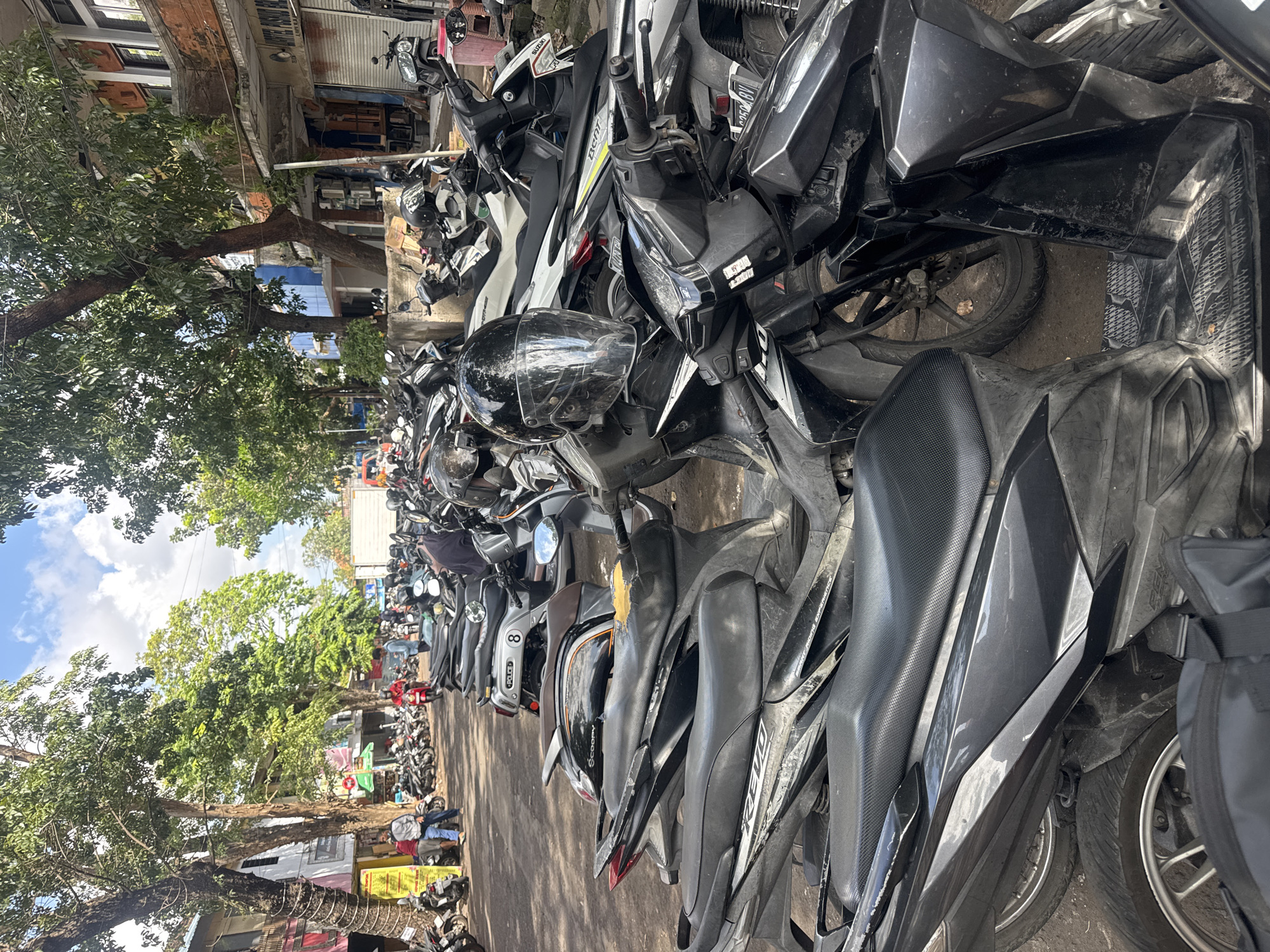 A street scene in Denpasar, Indonesia, is captured on a sunny day with a blue sky and scattered clouds. The foreground is dominated by a dense parking lot filled with numerous motorbikes, predominantly black and dark colored. Several motorbikes are parked closely together, with their seats and handlebars visible. One prominent black motorbike in the center has a clear-visored helmet resting on its handlebars. Further back, the street extends into the distance, lined with trees on both sides. On the right side of the street, buildings with balconies and shops are visible. On the left, there are more trees and a yellow signpost. A few people are scattered throughout the scene, some standing near the parked motorbikes and others walking along the street. The ground appears to be a mix of dirt and paved surfaces. The overall impression is of a busy urban area with abundant motorbike traffic and parking.