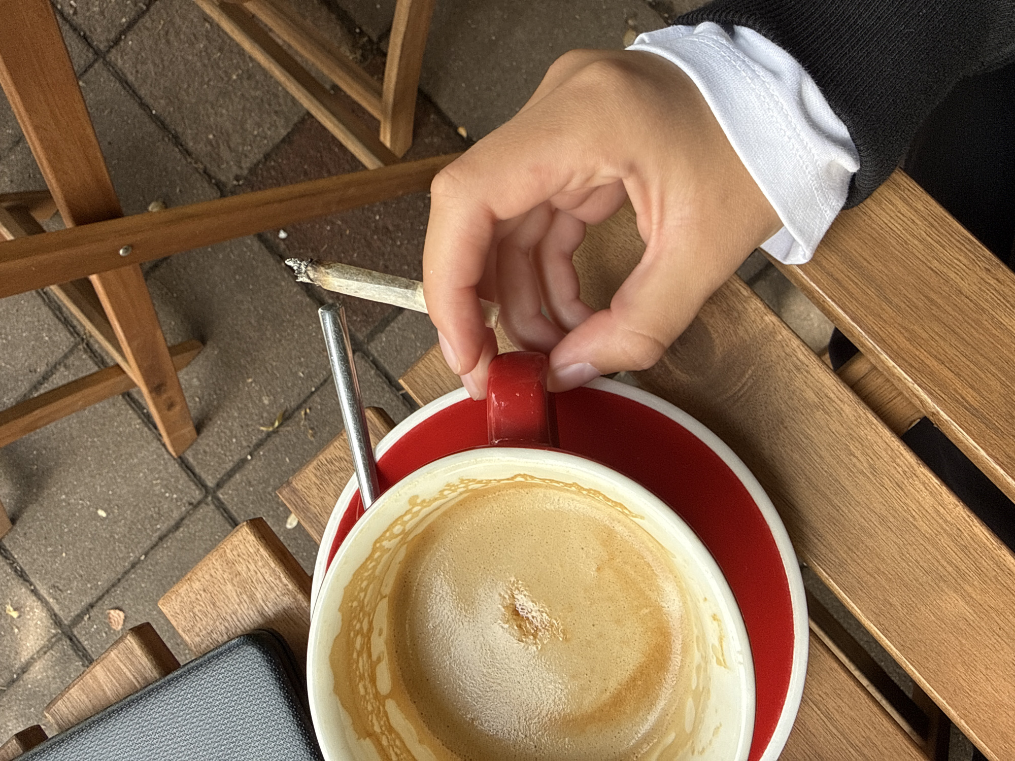 The image depicts a close-up, overhead view of a person's hand holding a cigarette, which is positioned near a cup of coffee and a metal stirrer on a wooden table. The coffee is light brown with foam. The cup is white with a red rim and sits on a matching red saucer. The hand is holding the handle of the cup. Behind the coffee, a portion of a dark-colored object, possibly a phone or tablet, is visible on the table. Wooden chairs and brick pavement are visible in the background, suggesting an outdoor seating area, potentially at a cafe in Belgrade, Serbia. The lighting suggests it is daytime.