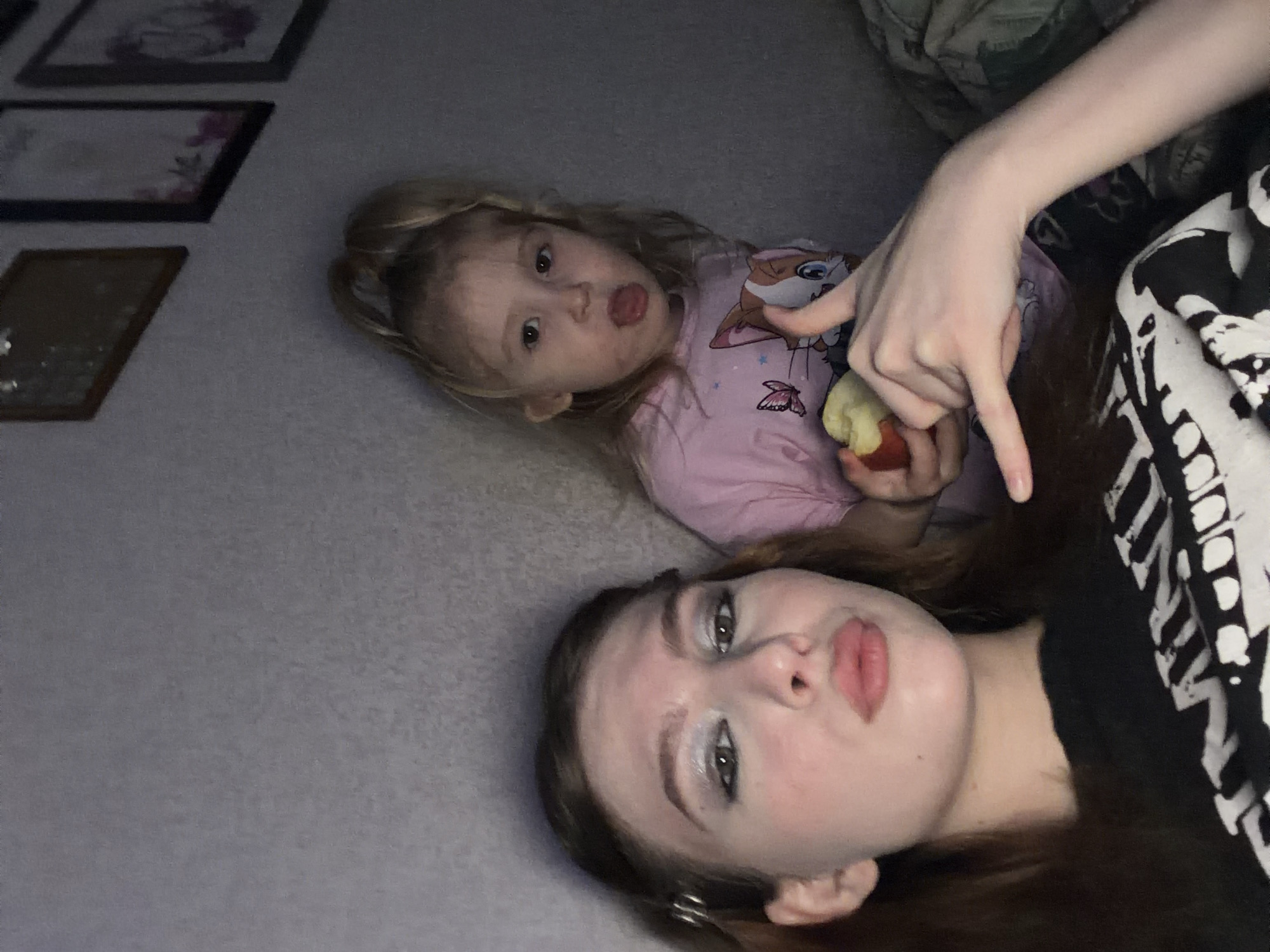 Two individuals, seemingly a young girl and an older teenager or young adult, are positioned on a soft surface, possibly a rug or blanket. The young girl, positioned at the upper left of the frame, wears a pink shirt with a cartoon character print and is holding an apple. She is sticking out her tongue towards the camera. An arm with a pale hand and elongated fingers is pointing down at the girl. The older individual lies closer to the bottom of the frame, with their face angled towards the viewer. They have dark hair and are wearing makeup with light eyeshadow. Their lips are pursed in a kissing expression. They are wearing a black shirt or garment with a white, patterned design.
Three framed pictures are visible on the upper left of the image. The background appears to be a simple, possibly indoor setting with minimal detail.
The image appears to be taken from a slightly elevated angle, possibly a selfie, with a tilted perspective. There is no discernible evidence to suggest the location is Noviy, Russia.