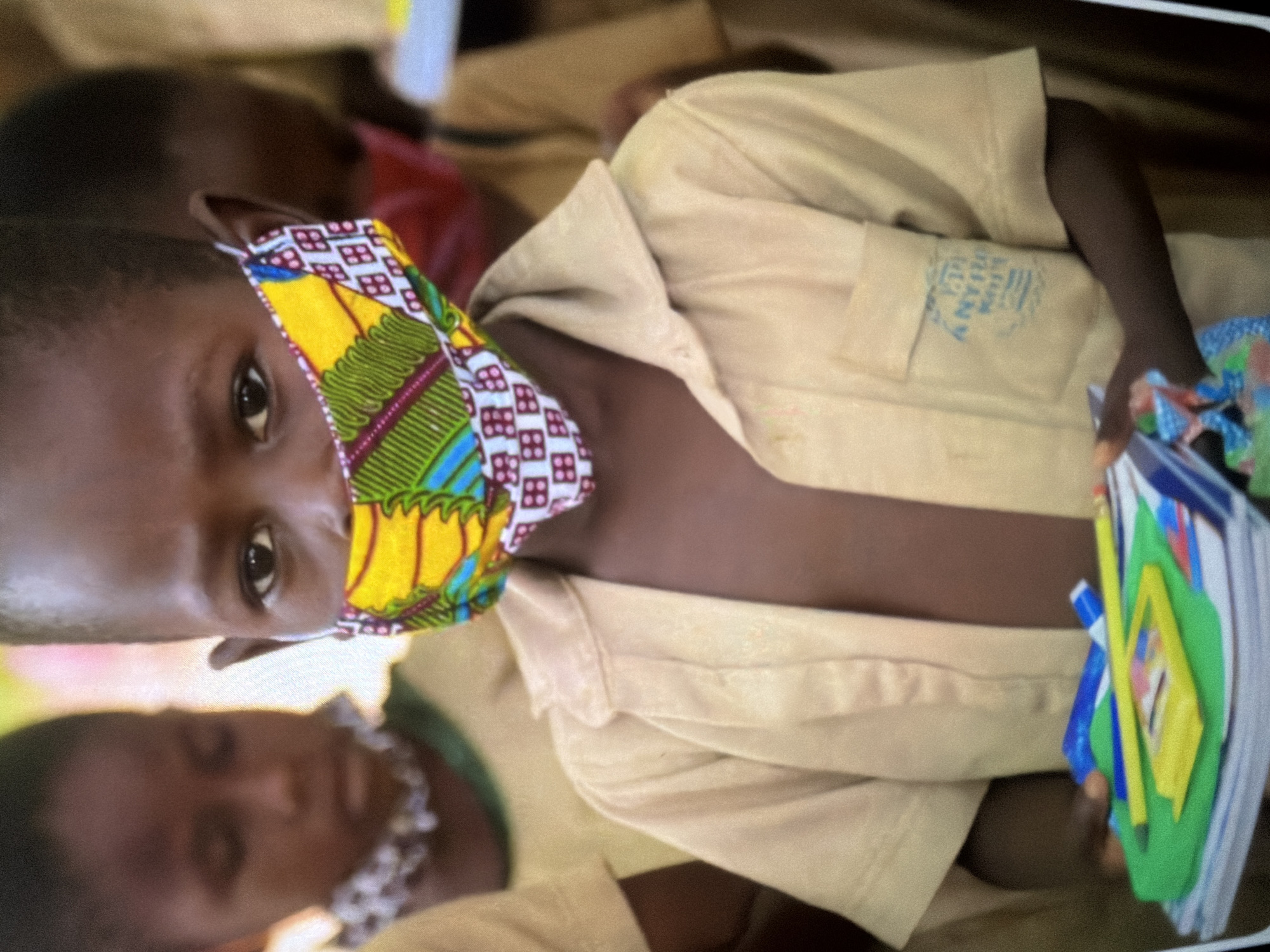 A close-up shot shows a young Black child wearing a tan collared shirt and a colorful face mask. The mask has a yellow base with green, blue, and red geometric patterns. The child's right eye is visible and looking towards the camera. In front of the child, a stack of notebooks is held, with a green folder on top. A yellow pencil and a blue pen are resting on the green folder. The child's hands are partially visible, holding the materials. Other children are blurred in the background, suggesting a school or outdoor gathering. The background is out of focus, with muted colors and a soft lighting. A logo is visible on the child's shirt pocket.