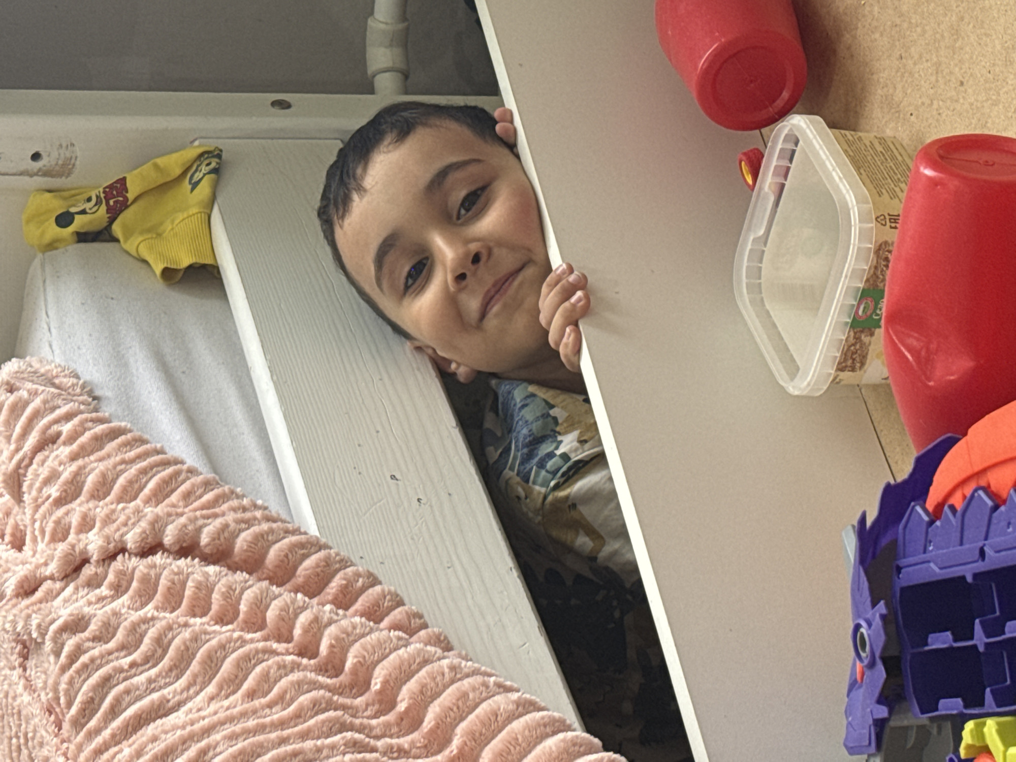 A young boy is peering out from between a white door or panel and a vertical wall. The boy has dark hair and eyes and a slight smile. He is wearing a shirt with a floral pattern. To the left, a white surface is partially covered by a peach-colored, textured blanket and a yellow sweatshirt with a cartoon character design. To the right of the boy, a white container, two red plastic cups, and a purple toy are visible. The environment appears to be a small, enclosed space, possibly a closet or a built-in storage area. There is no information to support the location being in the city of Chaikovsky, Russia.