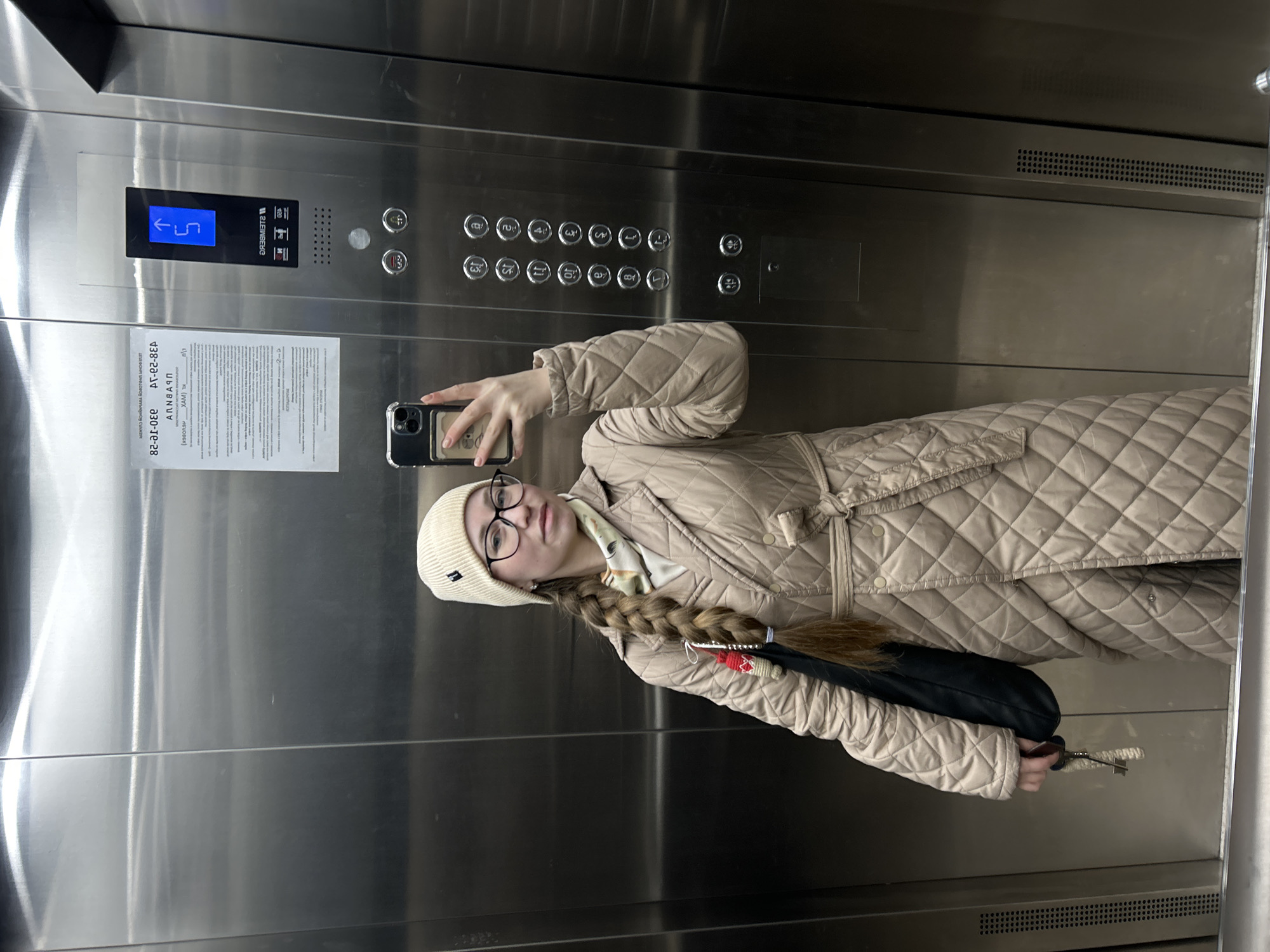 A young woman takes a mirror selfie inside a modern elevator. She stands centrally in the frame, wearing a light beige quilted coat, a matching beanie, and dark-rimmed glasses. Her long, thick brown hair is styled in a braid, adorned with red and white decorative ties. She holds a smartphone, visible in the reflection, to capture the image, with a dark handbag hanging from her right arm.
The elevator's interior features polished stainless steel walls. To her left, a control panel is visible, equipped with a digital display showing "5" with an upward arrow, indicating the elevator is on its way to the fifth floor. Below this, there are numerous silver buttons for floors 1 through 13, along with standard elevator controls for door open/close, an alarm bell, and a fan.
To the left of the control panel, a white paper notice is affixed to the wall. Partially legible text includes "PLEINBEKE," "ANNÄNN," and sequences that resemble contact numbers or codes, such as "438-22-14" and "230-те-28." Other text, including "XAM" and some Cyrillic characters, is also faintly visible. The woman's expression is neutral, consistent with posing for a photograph. The setting is an indoor location, an elevator within a building in Saint Petersburg, Russia.