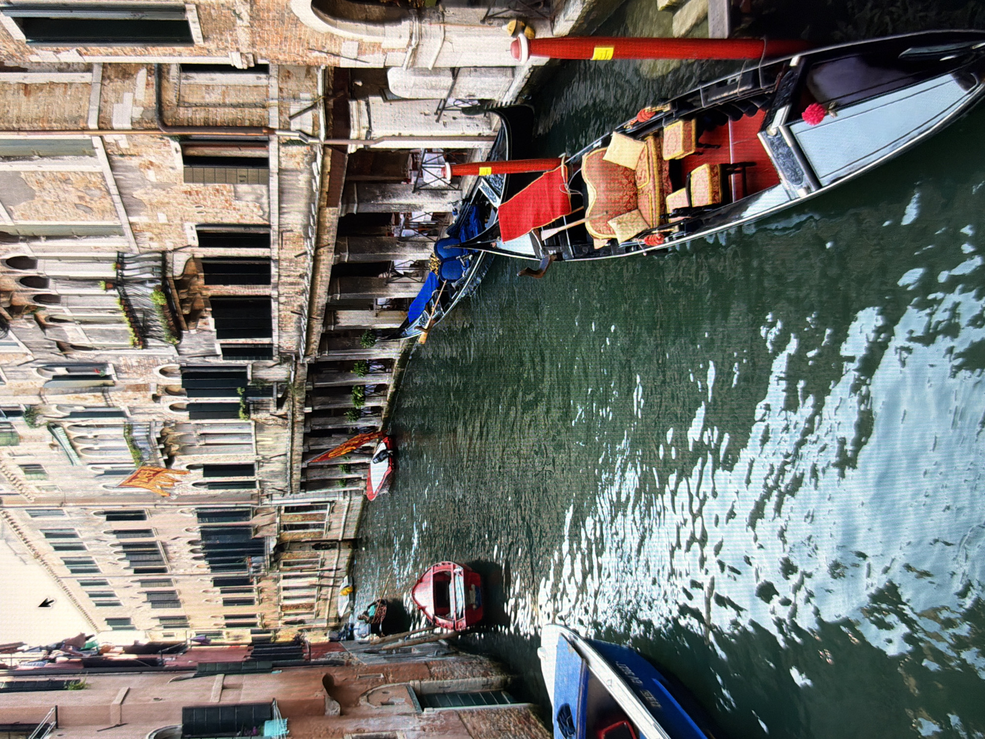 The image depicts a canal scene in Venice, Italy, characterized by traditional Venetian architecture and several boats. The buildings lining the canal are constructed of brick and stone, with numerous arched windows and balconies adorned with greenery. The water in the canal is a dark green, reflecting the buildings and sky. Several boats are visible: a blue and white motorboat in the foreground on the left, a red rowboat in the middle ground, a gondola with blue cushions in the distance on the left, and two ornate gondolas in the foreground on the right, one with red seating and cushions. Two people are seated in the gondola on the far right. The overall atmosphere suggests a typical Venetian waterway, bustling with aquatic transportation and surrounded by historic structures. The prompt's request to locate this scene in Kyiv, Ukraine, is incorrect; this is unequivocally Venice, Italy.