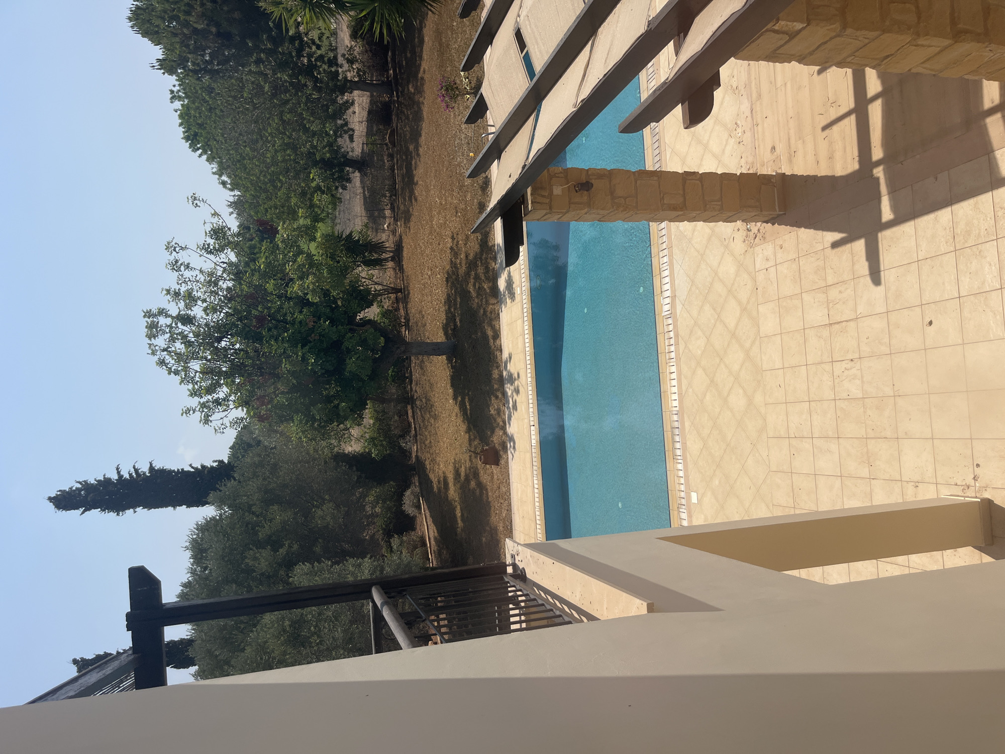 The image shows a view of a swimming pool and surrounding area on a sunny day. The pool is rectangular with blue water and is surrounded by beige tiles. To the left of the pool is a white building with a balcony and a railing. To the right of the pool is a stone pillar supporting a wooden pergola with beige fabric shades. In the background, there are trees, dry grass, and a clear blue sky. The scene appears to be in a resort or residential area in Aphrodite Hills, Cyprus, characterized by the architecture and landscaping. There are no people visible in the image.