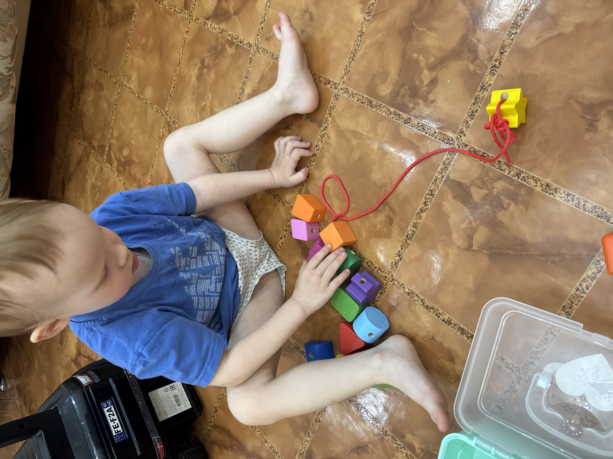 A child is sitting on a brown tiled floor in a room in Ферма, Russia. The child is wearing a blue shirt and light-colored shorts. The child is playing with colorful wooden blocks and a red string. A black toy car is to the left and a clear plastic container is on the right.