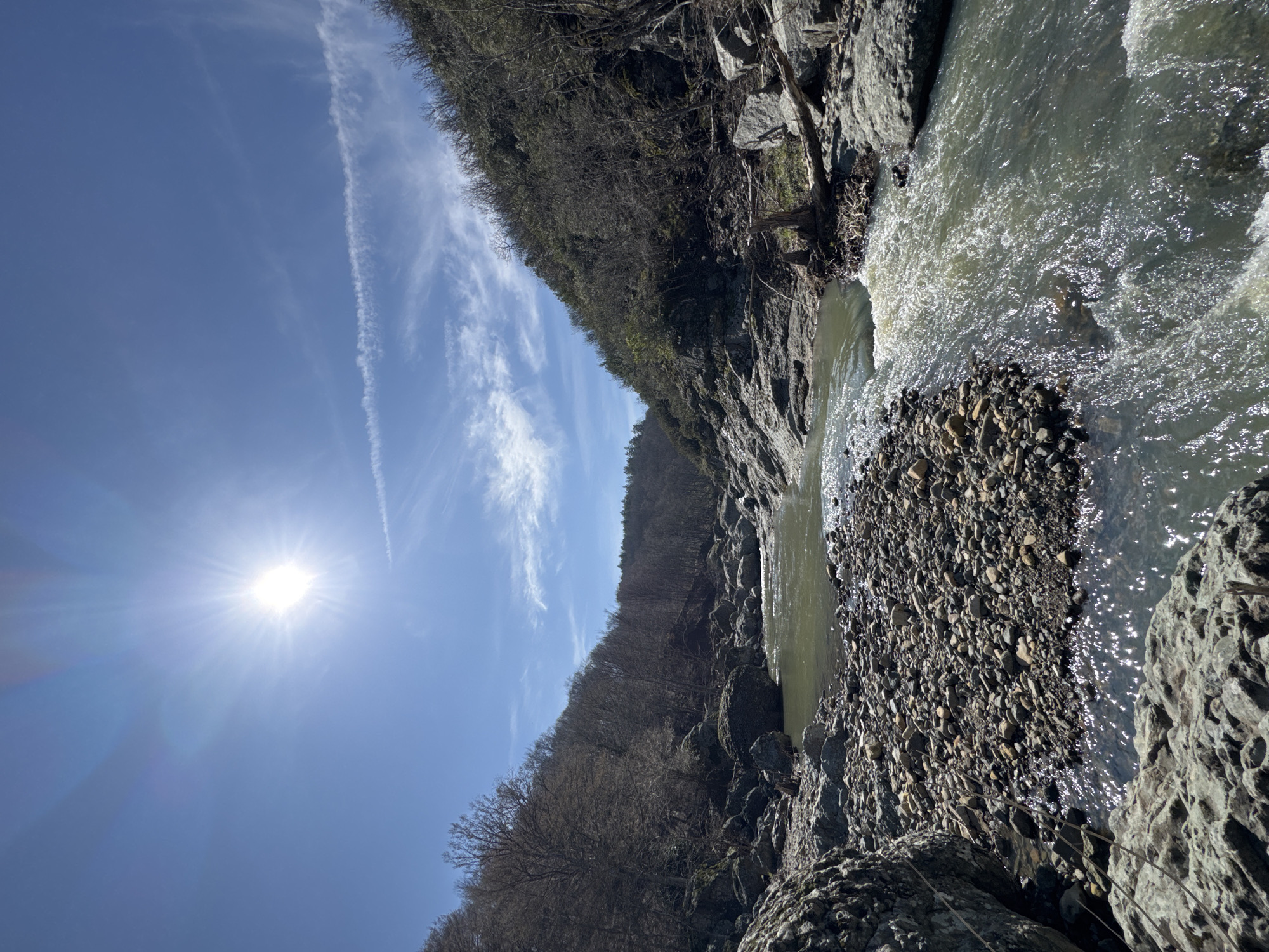 The image depicts a natural riverine landscape within a valley in Fazanovo, Bulgaria, under clear, bright daylight. The sun, positioned in the upper left quadrant, radiates intensely, creating lens flare and casting direct light on the scene. The sky is a vibrant blue with sparse, wispy cirrus clouds and faint contrails in the upper right.
A moderately wide river, characterized by murky greenish-brown water, flows from the background towards the foreground. Its surface displays significant turbulence and sparkling reflections of sunlight, particularly on the right side. The riverbed and banks are extensively rocky, composed of numerous grey and tan boulders, rocks, and smaller pebbles. A prominent, elongated shoal of assorted pebbles and gravel partially divides the river in the mid-ground.
Steep, forested hillsides flank both sides of the river, ascending sharply. The trees on these hillsides are predominantly deciduous, appearing bare of leaves, suggesting a late winter or early spring season. Some darker evergreen foliage is visible further up and in the distance. Scattered natural debris, including fallen logs and tree stumps, is present along the rocky riverbanks, notably on the right side of the frame. No human individuals, animals, or artificial structures are visible within the immediate area.