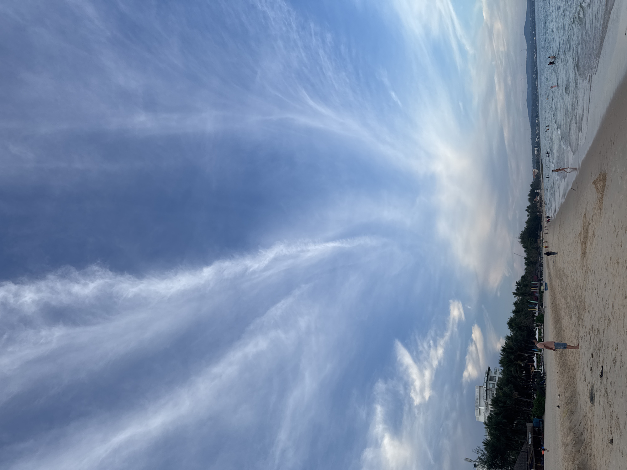 This is a vertical, wide shot of a beach scene under a vast, partly cloudy sky. The sky is a dominant feature, with wispy, white cirrus clouds streaking across a deep blue expanse. The sunlight filters through, creating bright areas near the horizon and a softer, diffused light higher up, suggesting either early morning or late afternoon.
On the right side of the frame, a sandy beach stretches along the coastline. Gentle waves lap at the shore of the ocean. Several people are visible, some walking along the beach, others in the shallow water. One person in the foreground on the beach is wearing blue shorts and a light-colored top. Further down the beach, a line of trees and buildings suggests a developed area or resort. In the distance, a mountainous or hilly landscape can be seen along the horizon. The overall atmosphere appears relaxed and leisurely, typical of a beach day.
The scene is likely taking place in Phu Thuy Ward, Vietnam, given the location context. There is no visible text in the image.