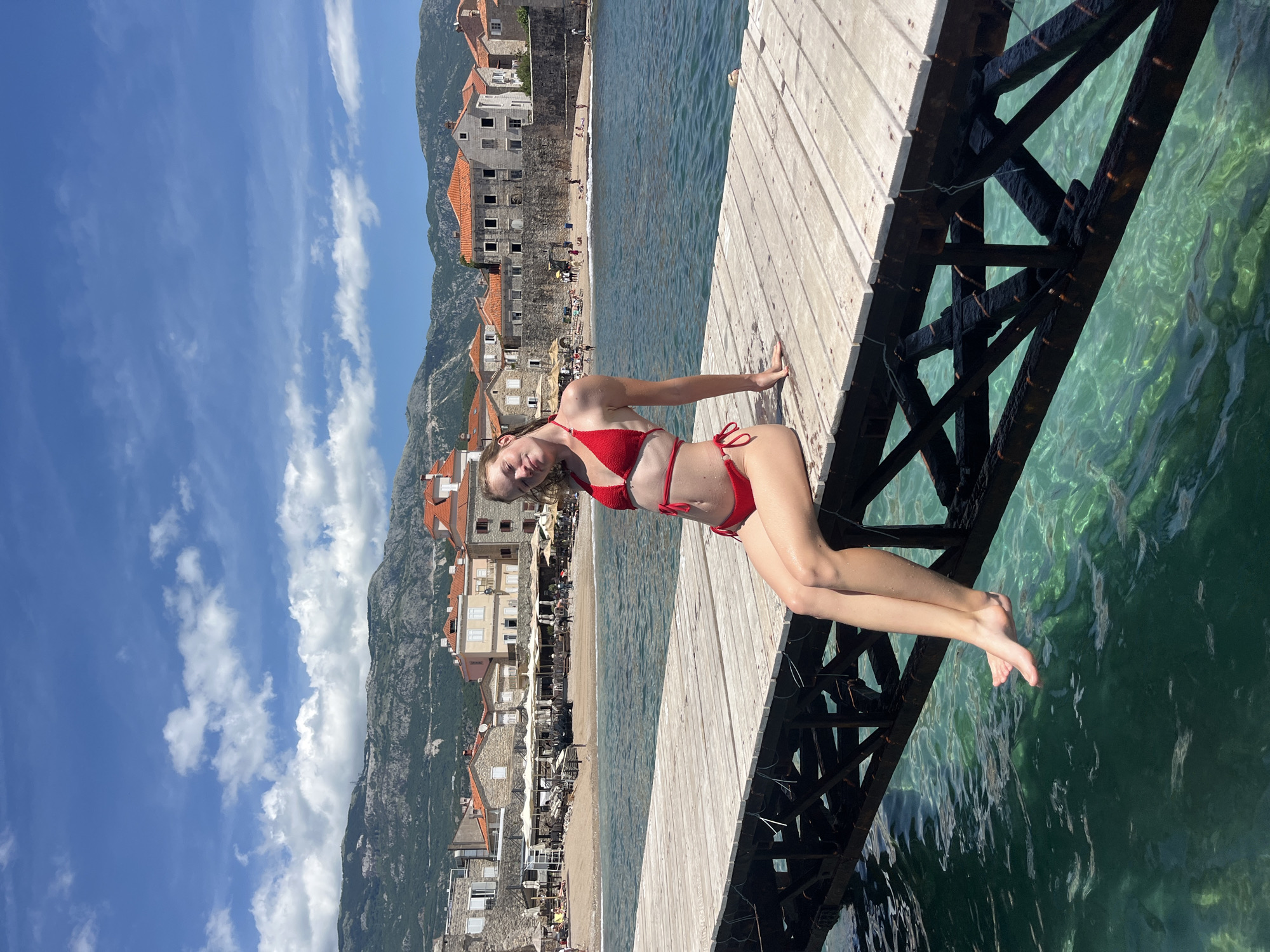 The image depicts a coastal scene on a bright, sunny day, consistent with the city of Budva, Montenegro. In the foreground, a young female individual, appearing to be in her late teens or early twenties, is seated on a weathered wooden pier. She is positioned towards the left side of the frame, wearing a red two-piece bikini, with her bare legs dangling over the clear, turquoise-green water but not submerged. Her right hand rests on the pier for support, and her eyes appear closed or mostly closed, indicating a relaxed posture. Small fish are discernible in the transparent water beneath the pier, which is supported by a dark, possibly metal framework showing signs of wear.
The midground features a sandy beach to the left and right of the pier, populated by numerous individuals, sun loungers, and beach umbrellas. Behind the beach, a collection of two-to-three-story buildings with light-colored facades and distinct terracotta-tiled roofs characterize a coastal town. Some buildings exhibit visible stone masonry. Outdoor dining areas with additional umbrellas are observable along the beachfront.
In the background, a range of large, rocky mountains covered with sparse vegetation stretches across the horizon under a bright blue sky dotted with scattered white cumulus clouds. The overall scene captures a vibrant summer atmosphere in a Mediterranean coastal setting.