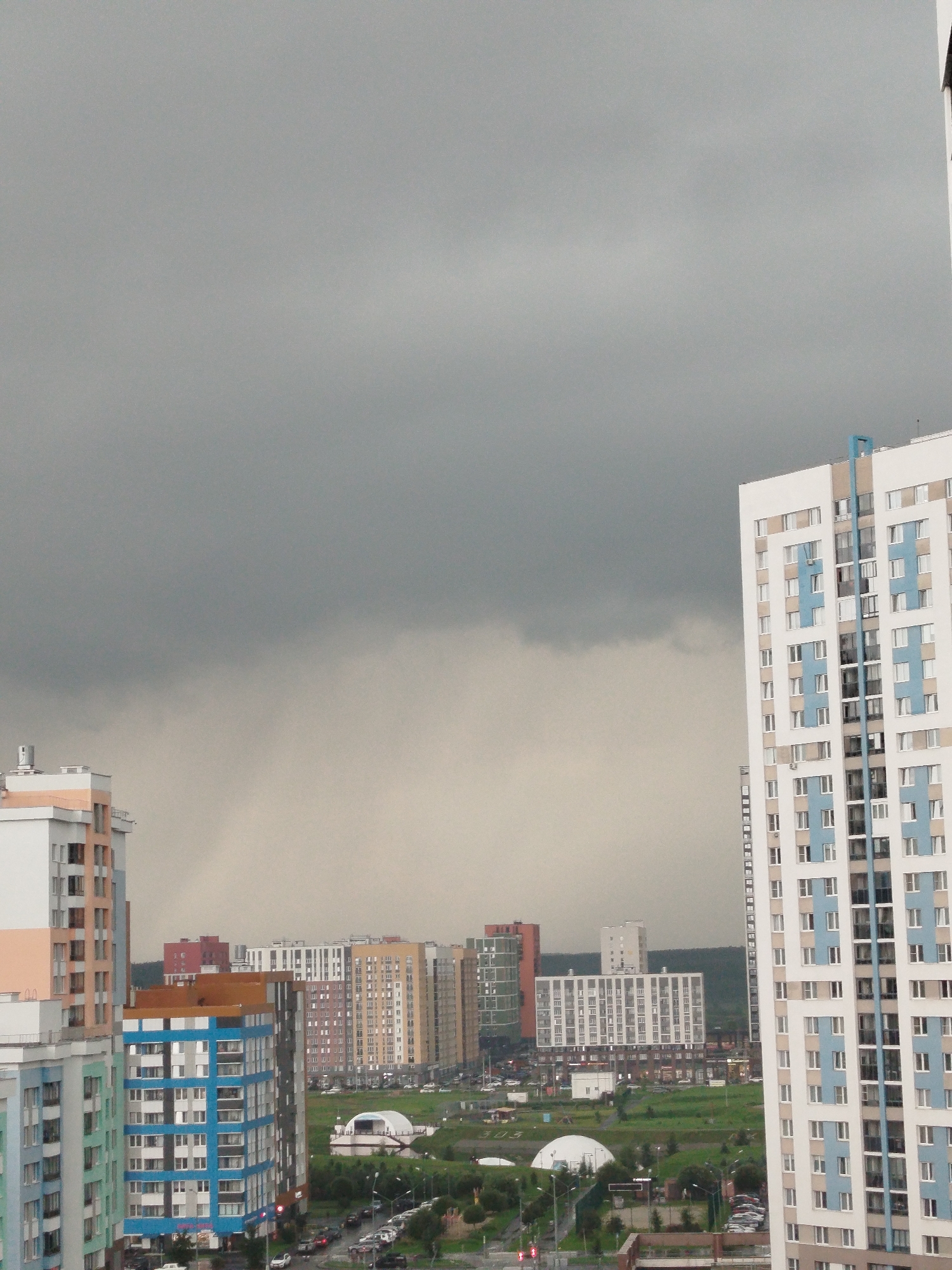 This is an aerial view of a dense urban residential area in Yekaterinburg, Russia, under a heavily overcast sky, suggesting impending or ongoing precipitation. The foreground is dominated by a tall, modern, white and blue apartment building with numerous windows. To the left, a slightly shorter apartment building with light orange and white facades is partially visible. In the middle ground, a cluster of similarly modern, multi-story apartment buildings of varying colors and architectural styles (including white, beige, brown, and red) form a cityscape. The buildings exhibit repetitive window patterns typical of contemporary housing developments. A white, dome-shaped structure, possibly a sports facility or exhibition hall, is visible in the mid-ground amidst green landscaping and a network of roads. The roads are lined with parked cars, indicating a populated area. The background features rolling green hills and a dark, oppressive cloud bank that covers most of the sky, creating a dramatic and somewhat somber atmosphere. No people are clearly discernible in the image.