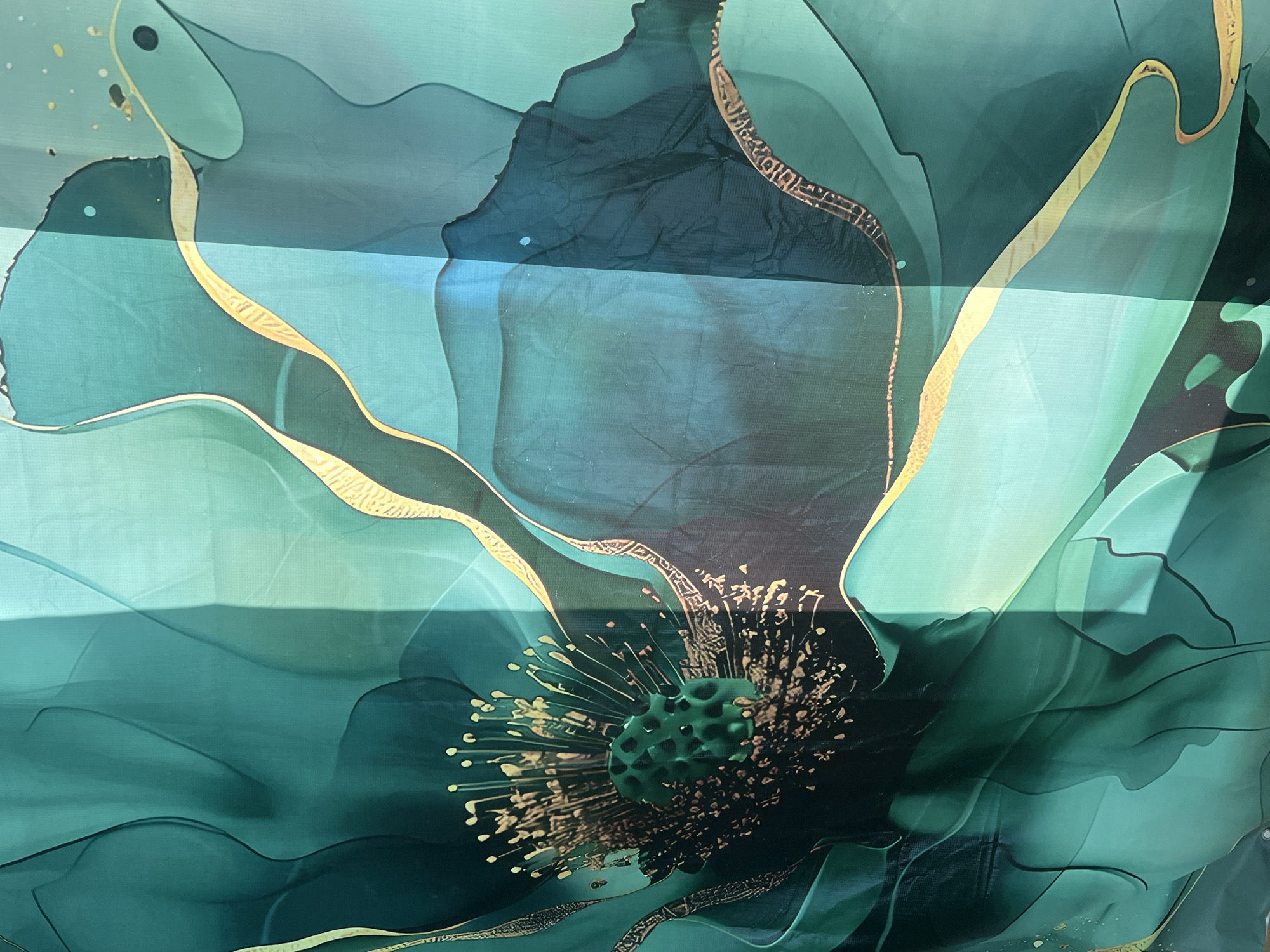 The image presents a close-up of a vibrant, abstract artwork or textile print, dominated by a large, stylized flower. The primary colors are a rich palette of teal, emerald green, and deep blue-green, creating a fluid and organic aesthetic that evokes alcohol ink or watercolor techniques.
The central subject is the intricate core of the flower, which features a textured, dark green, possibly bulbous center resembling a seed pod. Radiating outwards from this core are numerous fine lines and dots, rendered in metallic gold and lighter green, suggesting stamens. Metallic gold accents also delineate the contours of many petals, providing a shimmering contrast against the cool green hues. These gold lines often incorporate delicate, abstract patterns.
The petals are depicted with sweeping, overlapping forms, contributing to a sense of depth and movement. Areas of the design vary in intensity, from translucent lighter greens to more opaque, deeper shades. The surface of the media, likely fabric or a canvas, exhibits a subtle weave or texture. Several horizontal bands of bright light traverse the image, suggesting reflections from an external light source, possibly filtering through window blinds. The scene is static, showcasing an artistic representation rather than an ongoing event. No text is visible within the image.