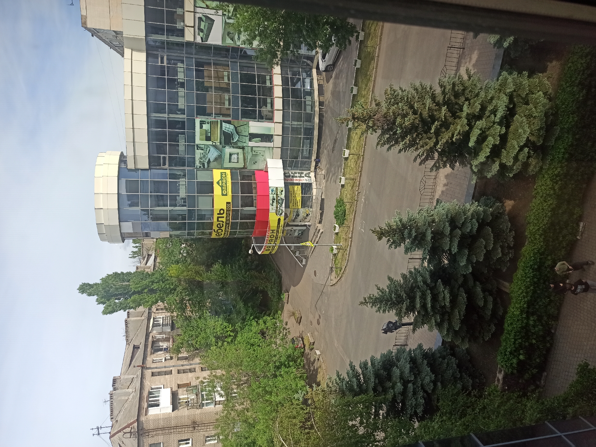 The image depicts an urban environment, possibly a street view in the city of Unknown, Unknown. A modern building with a glass facade is prominent, displaying advertisements or storefront signs. The building appears to be multi-storied. In front of the modern building, there are trees and shrubs. A road curves around the building. Further away, there is an older brick building with windows. On the sidewalk near the trees, several people are walking. The view is from an elevated position, possibly from a window. The weather seems fair.