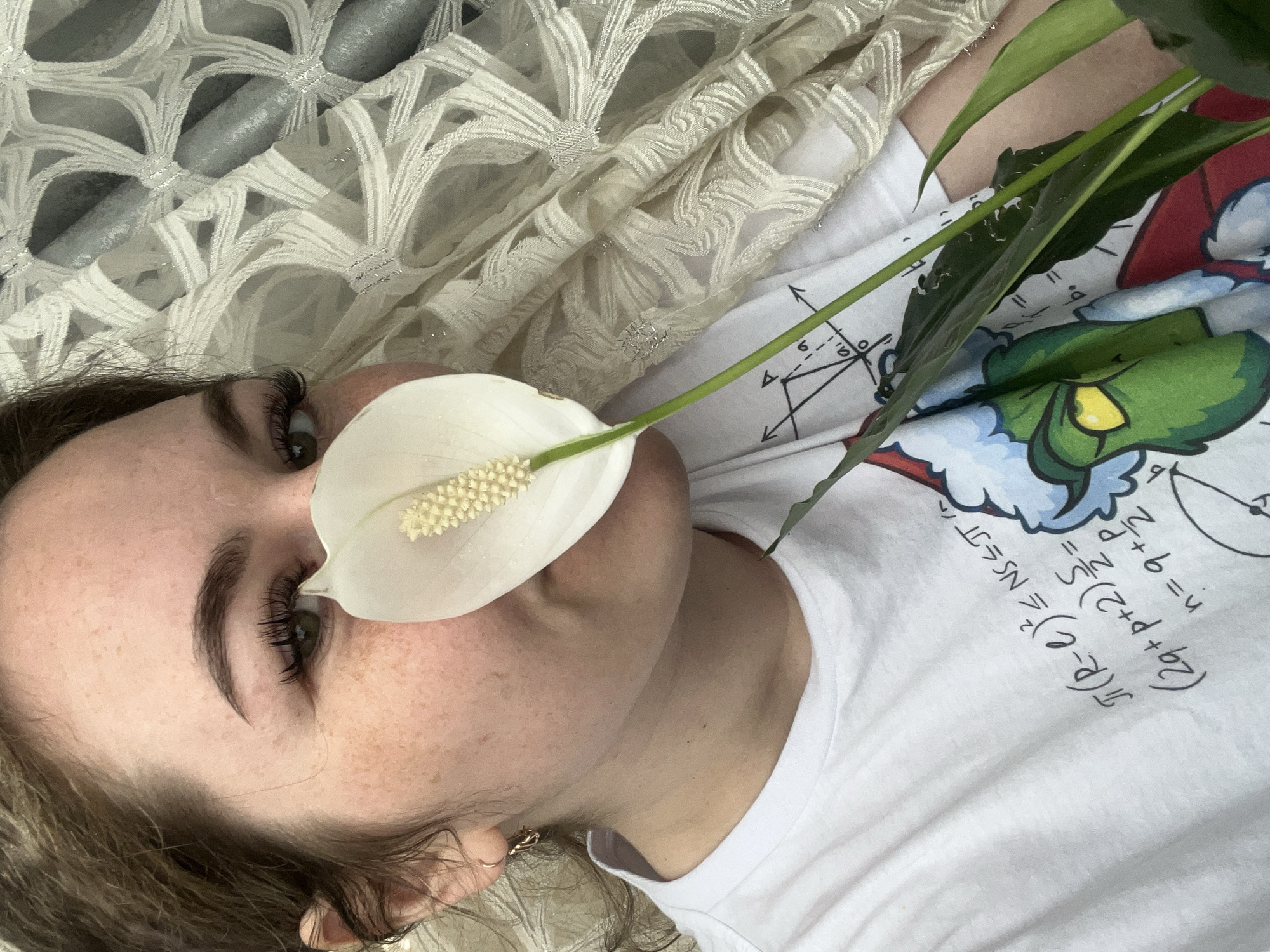 A woman is lying down. A white flower is placed near her nose. She is wearing a white t-shirt with mathematical equations and a cartoon of a character in the lower right of the shirt. A peace lily plant is near her. A lacy white fabric is above her. This image does not provide enough information to determine the location as Chaikovsky, Russia.