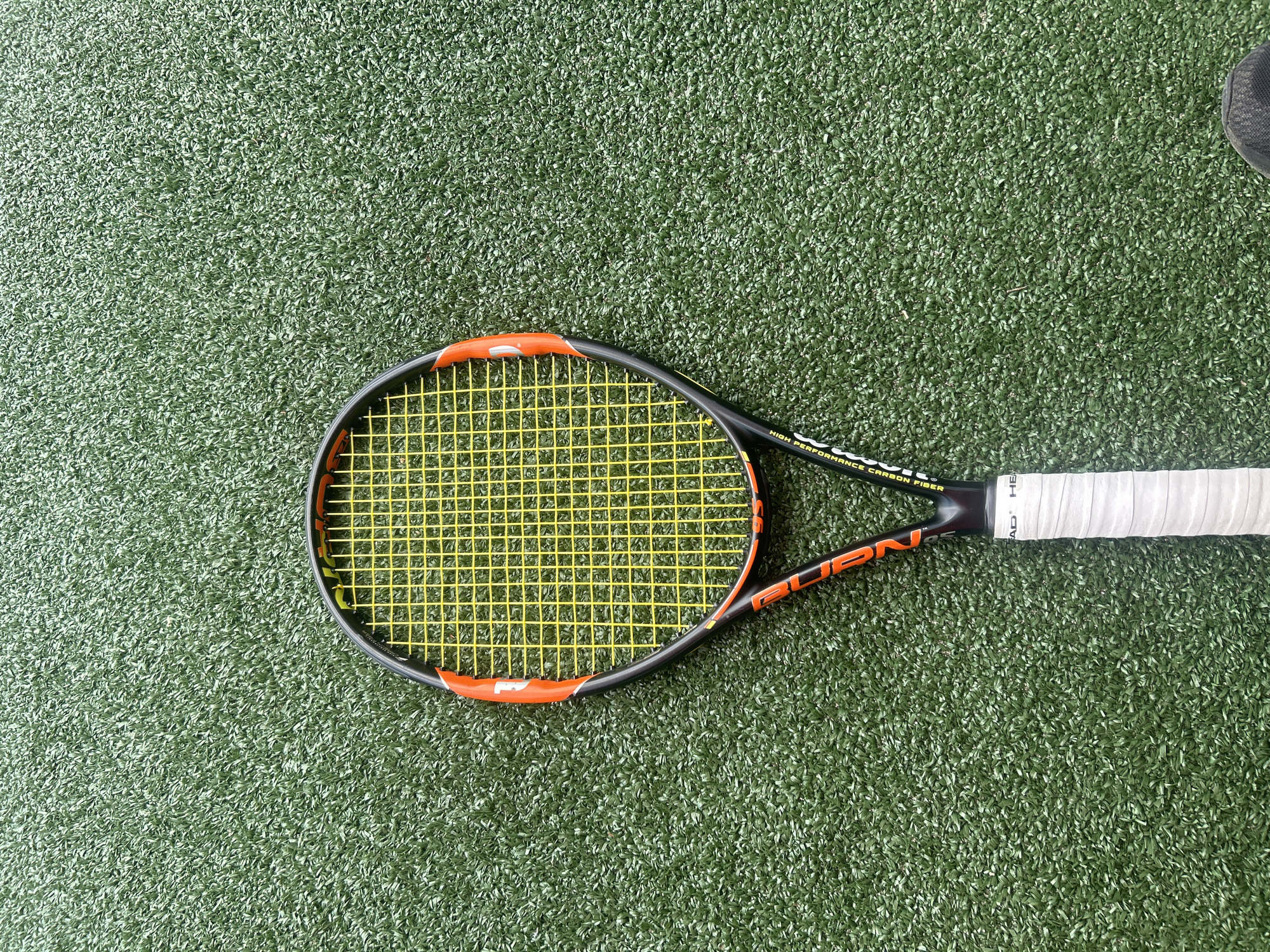 A close-up shot features a black and orange Wilson Burn tennis racket with yellow strings, lying on a green, artificial turf surface. The racket handle is wrapped in white tape. To the upper right of the racket, a portion of a black shoe is visible. The image does not show any recognizable landmarks to determine if the location is Veliko Tarnovo, Bulgaria.