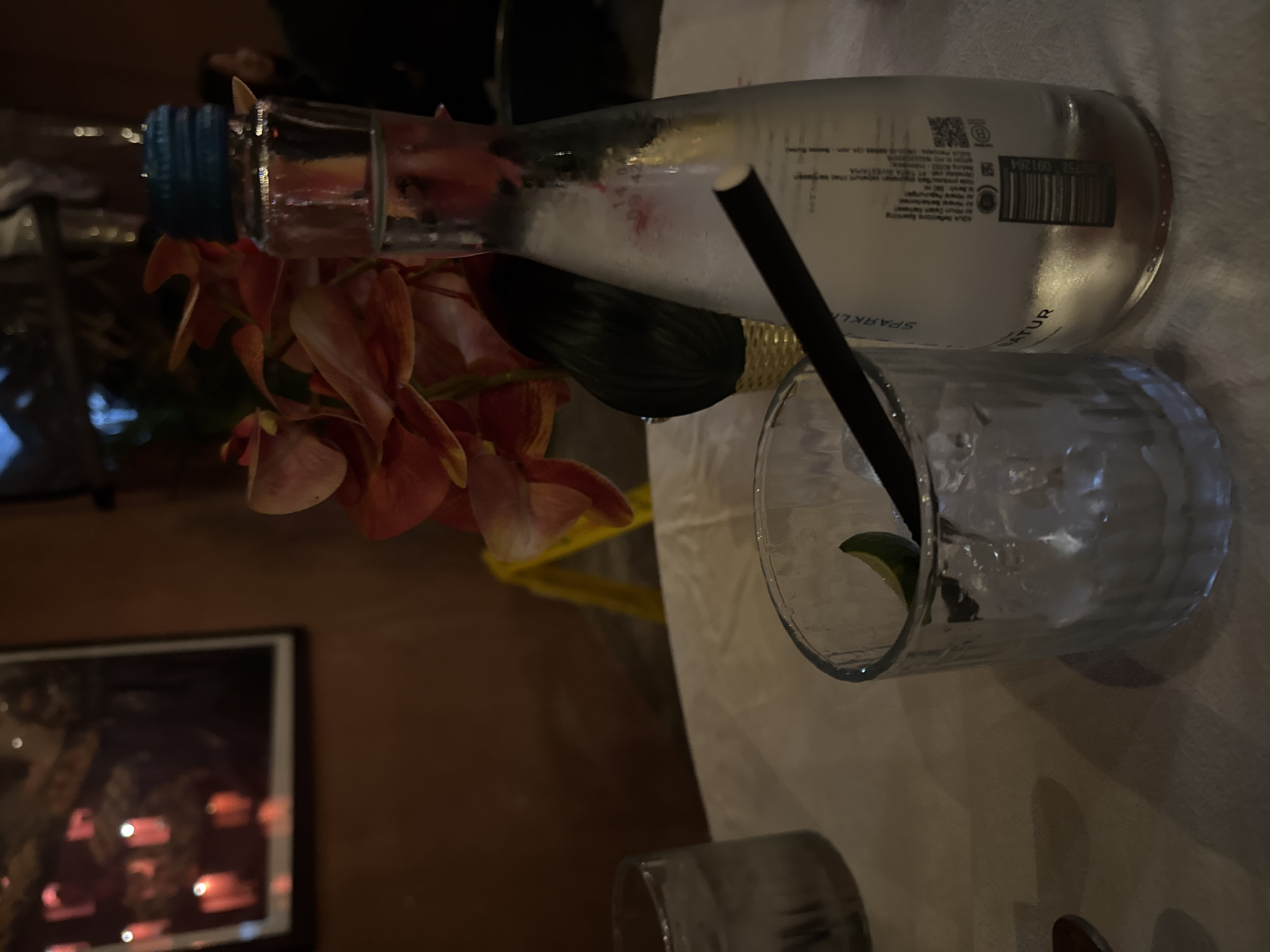 In Kuta, Indonesia, on a table covered with a white tablecloth, two glasses of ice are visible. One glass, on the left, is partially filled with a clear liquid. The second glass, on the right, contains ice and a slice of lime, with a black straw inserted. Next to the second glass is a glass bottle of sparkling water, its label indicating "SPARKLING NATUR." In the background, orange and red flowers are arranged in a woven vase, and a framed picture with red candle-like lights hangs on an orange-brown wall. The lighting is dim, suggesting an indoor setting like a restaurant or bar.