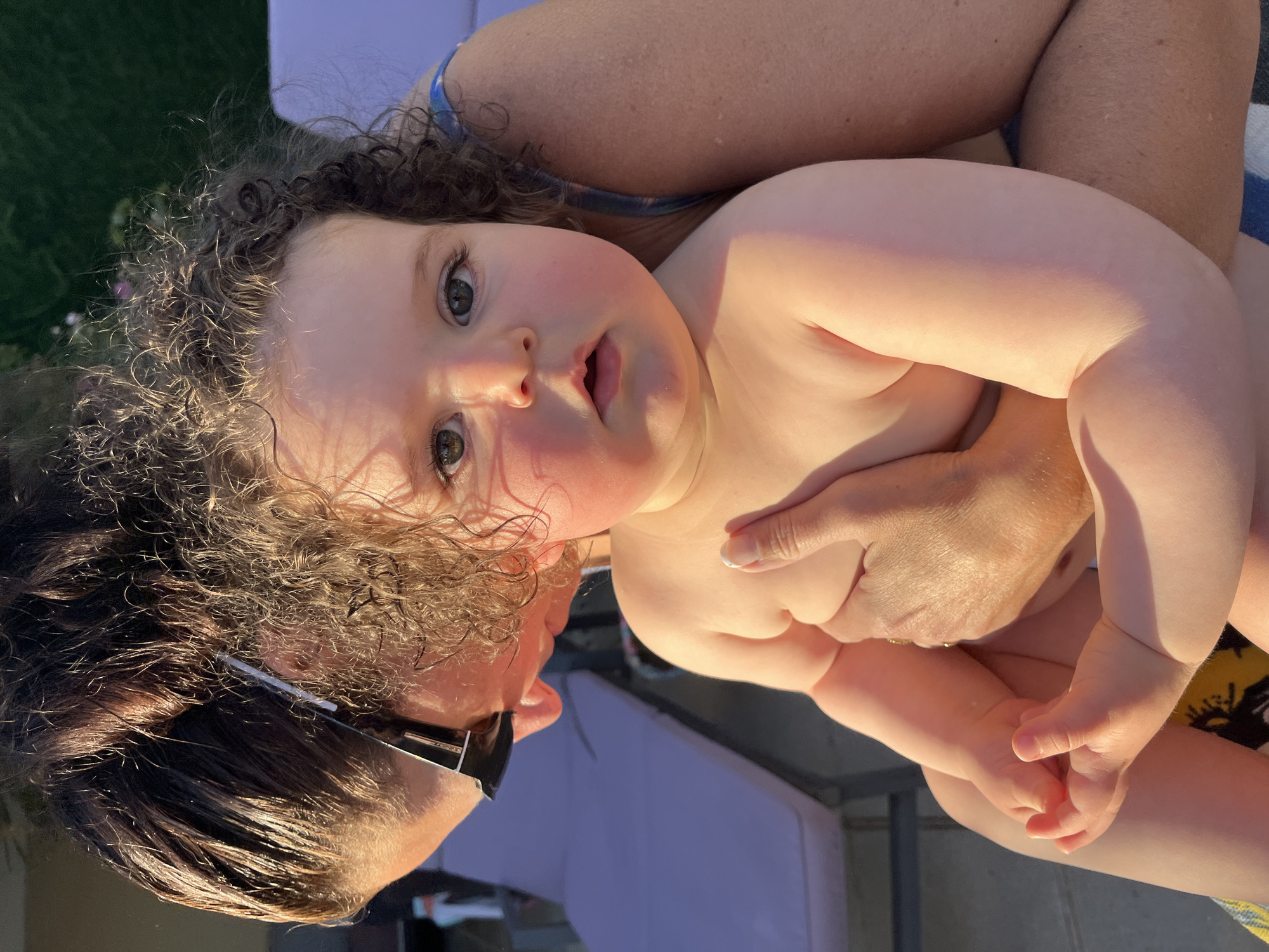 A close-up, low-angle, outdoor, natural light portrait shows a baby being held by an adult. The baby, in the foreground, has curly brown hair, wide, dark eyes, and rosy cheeks, with its mouth slightly open. The adult, in the background and out of focus, is wearing sunglasses and is holding the baby. The scene appears to be set in a backyard or patio area, possibly near a swimming pool, suggested by lounge chairs visible in the background. The overall lighting suggests it is late afternoon or early evening. The image's metadata does not provide specific location details within Saint-Galmier, France.