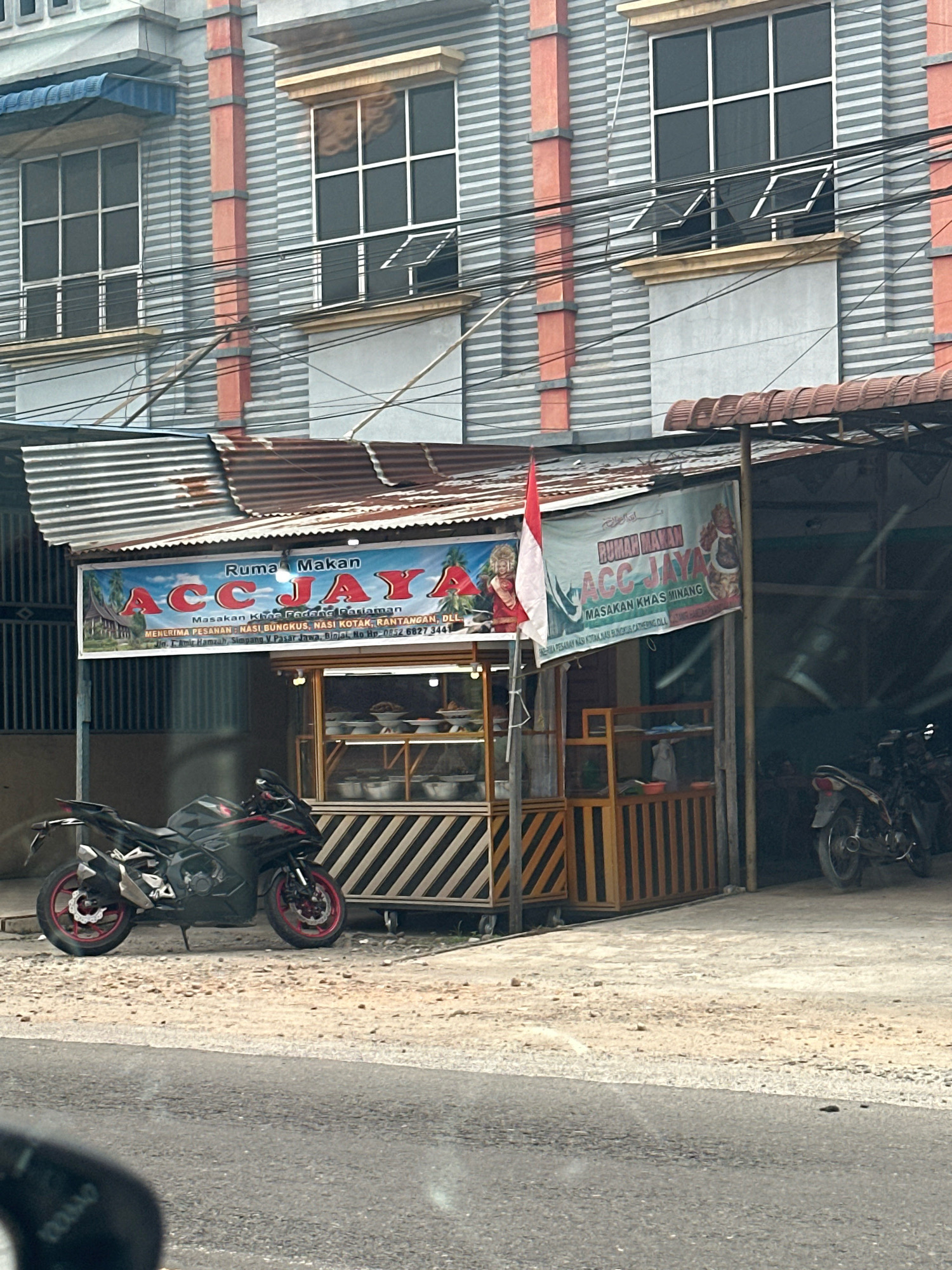 A street-level view captures a roadside eatery with a prominent banner in the city of Binjai, Indonesia. The eatery, identified as "Ruma Makan ACC JAYA," displays "Masakan Khas Padang Pariaman" and "MASAKAN KHAS MINANG" on its signage. It advertises catering services for "NASI BUNGKUS," "NASI KOTAK," and "RANTANGAN, DLL." The banner also provides an address: "Jl. T. Amir Hamzah, Simpang V Pasar Jawa, Binjai," and a phone number, "Hp. 0852 6827 3441."
In front of the eatery, two motorcycles are parked on a gravelly surface. One is a black sportbike with red accents, positioned on the left side of the frame. The other, a lighter colored motorcycle, is partially visible on the right. The eatery itself is a small, open-air structure with a corrugated metal roof, displaying food items in glass cases. Behind the eatery, a multi-story building with gray and orange striped facade and large windows is visible. Electrical wires are strung across the scene. The overall environment appears to be an urban setting in Binjai, characterized by its commercial establishments and street infrastructure.