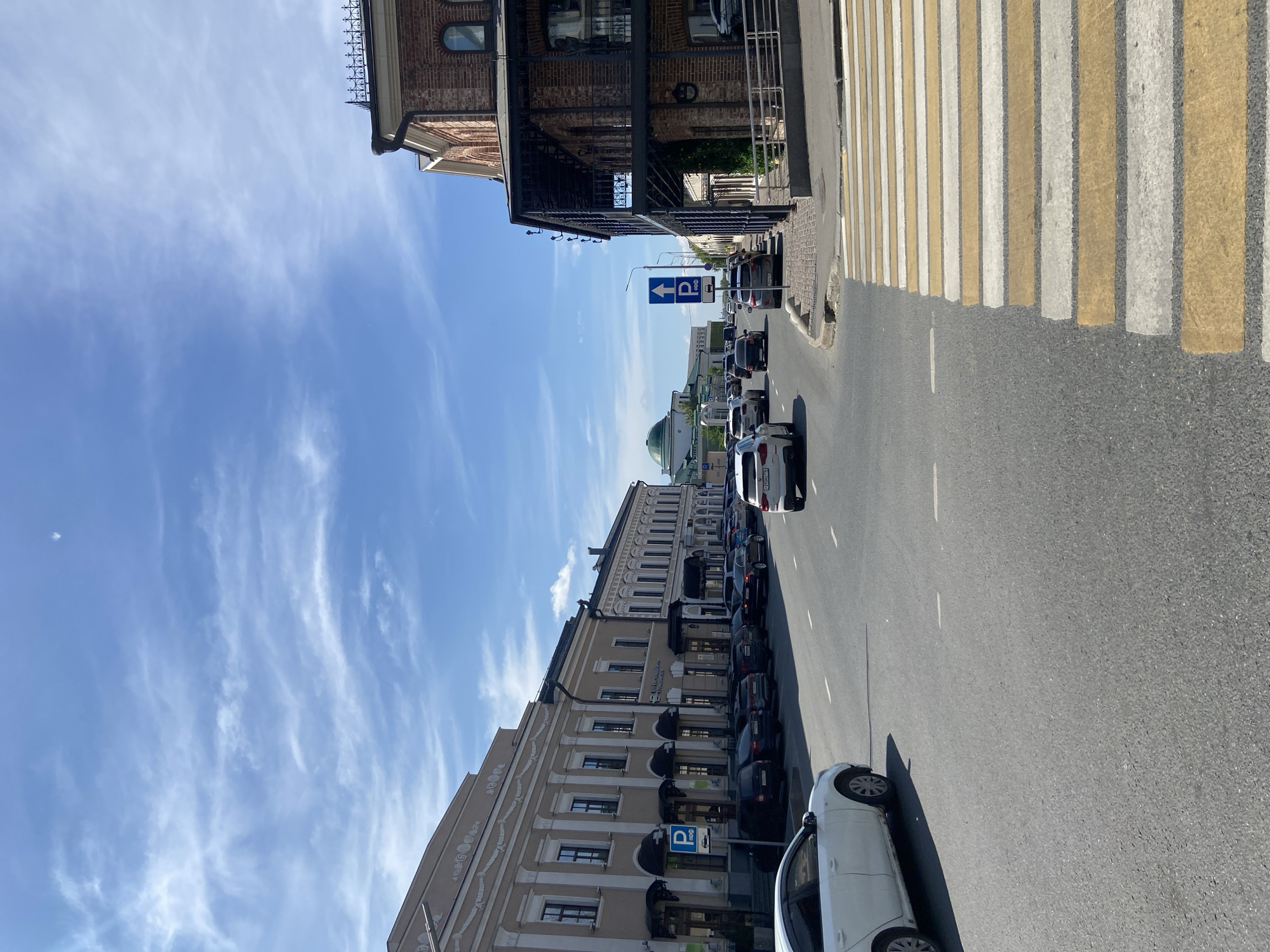 This image captures a wide-angle, sunny daytime street scene on Universitetskaya Street in Kazan, Russia, looking northeast towards the Kazan Kremlin. In the immediate foreground, a pedestrian crosswalk with alternating yellow and white stripes extends from the bottom right towards the center of the asphalt roadway. A white sedan is partially visible in the bottom left corner.
Along the left side of the street, a long, continuous structure of multi-story buildings with classical architectural elements, painted in a light beige hue, features numerous rectangular windows, ground-floor commercial entrances with dark awnings, and several parked dark-colored sedans and SUVs. A blue "P" parking sign with a downward-pointing white arrow is affixed to a pole on the far left.
On the right side, a prominent two-story building constructed of reddish-brown brick features arched windows, a substantial dark metal balcony structure on the first floor, and an ornate dark metal railing along its roofline. A small, cobbled sidewalk separates this building from the street. Further down the street, multiple vehicles are visible, including a white BMW X3 (Russian license plate "P210HA 116") on the right. A blue "P" parking sign with an upward-pointing white arrow and a paid parking symbol is mounted on a pole near the center-right.
In the distant background, slightly left of the central axis, the distinctive green dome of the Qolşärif Mosque, situated within the Kazan Kremlin, is clearly visible against a bright blue sky with scattered wispy clouds. No individuals are depicted within the frame.