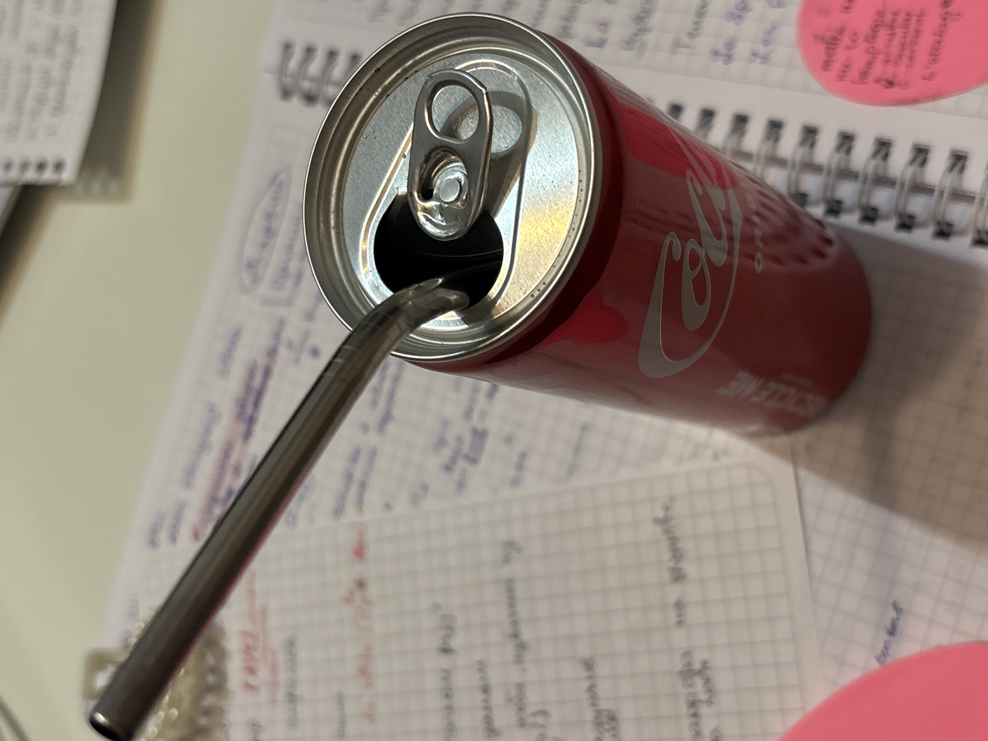 A close-up shot depicts an open red Coca-Cola can with a silver straw inserted, sitting atop a notebook. The notebook appears to contain handwritten notes and a pink circular sticky note is also visible. The scene suggests a setting where someone is possibly studying or working, with a drink nearby. The image provides no explicit indicators to pinpoint the location within Kragujevac, Serbia.