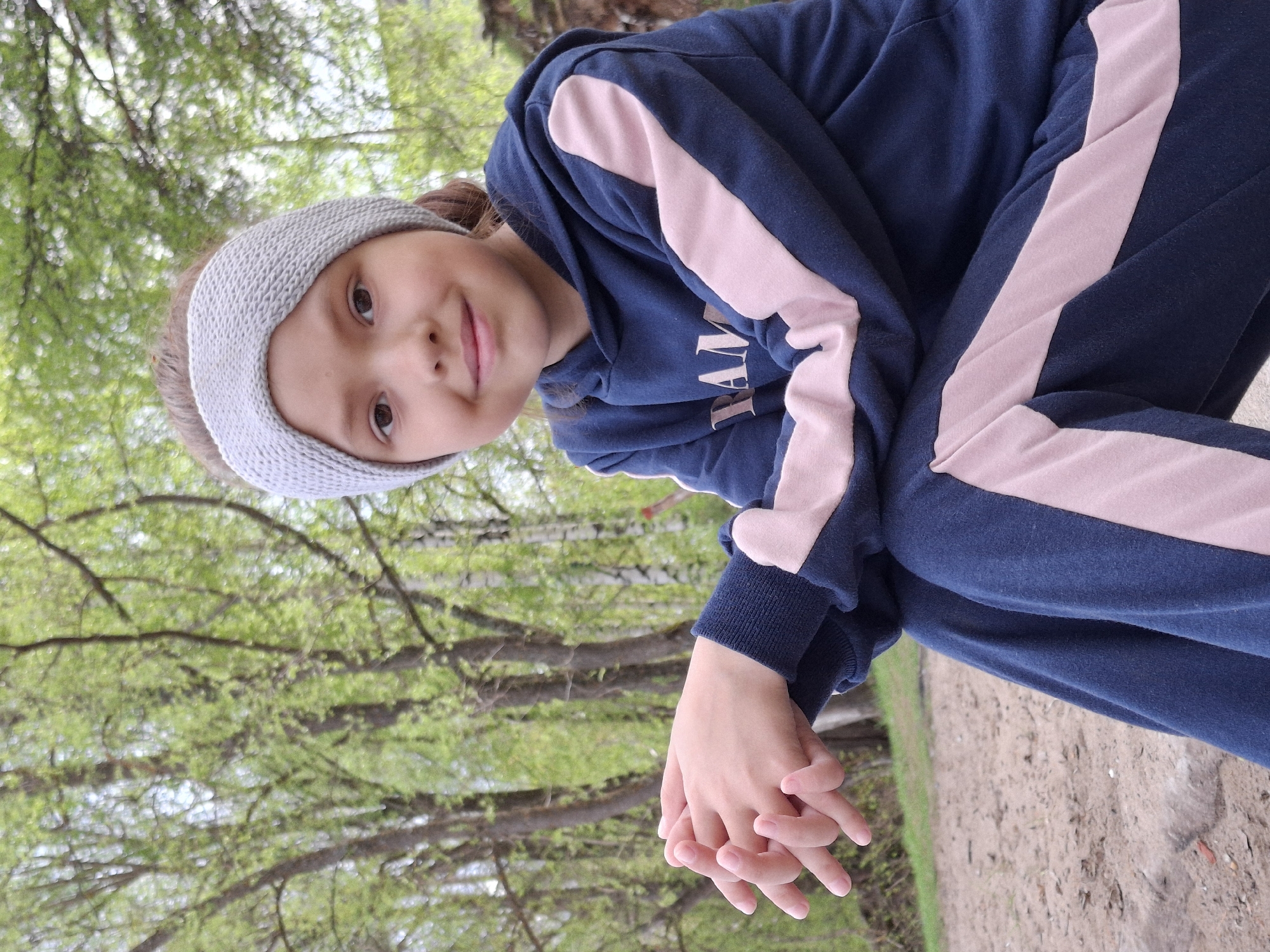 A young girl wearing a blue hoodie with pink stripes and a grey knitted headband is posing for a picture. She is holding her hands together in front of her. The background shows a forest-like area with green trees. A path or road is visible in the lower right corner. The location appears to be outdoors, possibly a park.