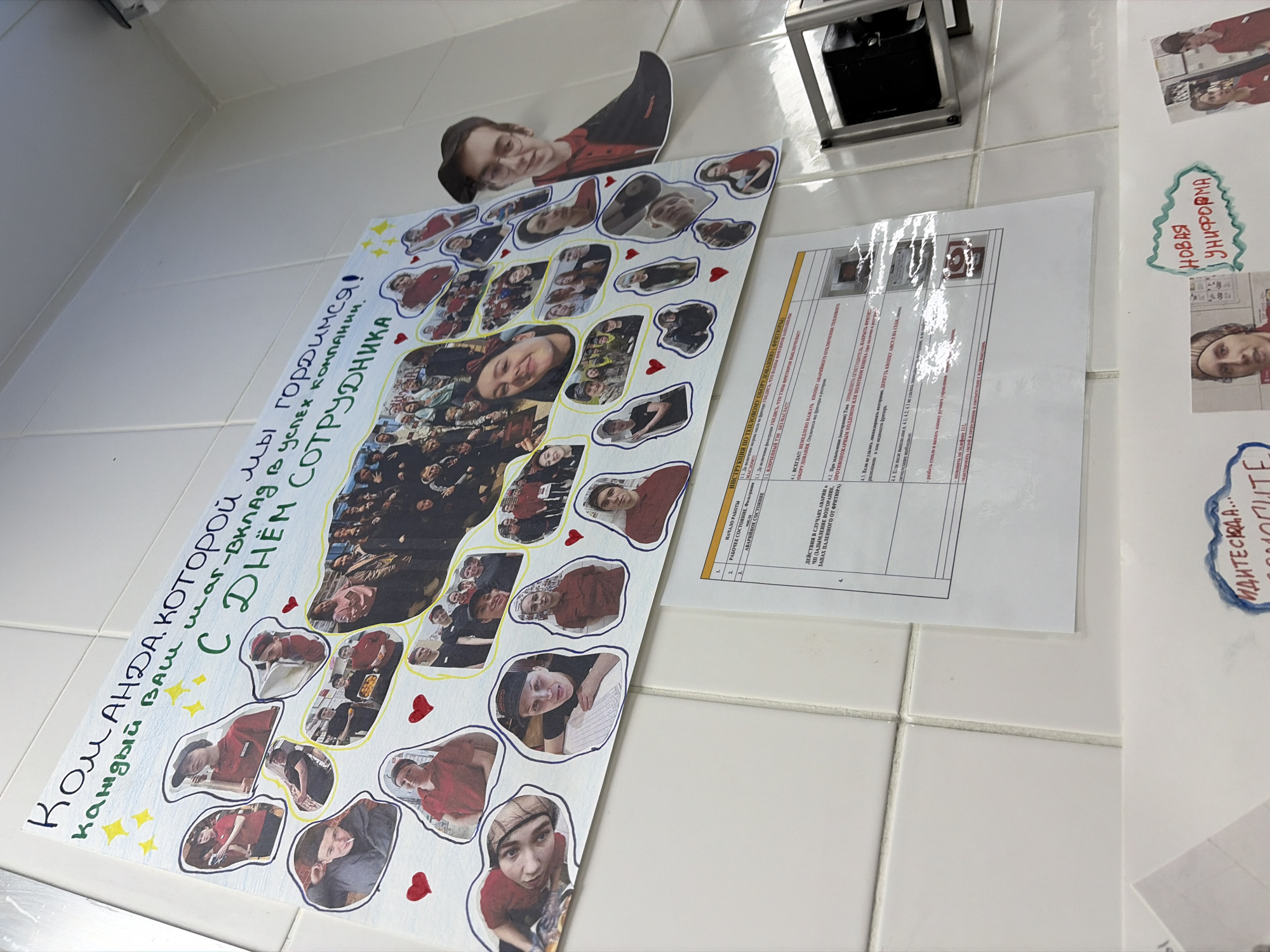 An indoor scene displays a white tiled wall in a commercial establishment within Chaikovsky, Russia. Dominating the upper center is a large, handmade collage poster celebrating "Happy Employee Day," featuring the Russian text "Команда, которой мы гордимся! каждый ваш шаг-вклад в успех компании. С Днём сотрудника!" (The team we are proud of! Every one of your steps is a contribution to the company's success. Happy Employee Day!). The poster is adorned with numerous cut-out photographs of individuals, predominantly young adults, often depicted in red or dark work uniforms, smiling and engaged in various activities, with a large central group photo and smaller individual portraits. Decorative red hearts and yellow stars are drawn around the photos. A three-dimensional cutout of a man in glasses and a hat wearing a red shirt extends from the right side of the poster. Below this main display, a white instruction sheet with numbered points and small photographic illustrations outlines procedures, including sections for "START OF WORK," "WORKING CONDITION," and "EMERGENCY CONDITION," providing instructions for actions in case of emergencies involving thermal equipment or frying. To the right of the instruction sheet, a metallic bracket or fixture holds a dark, rectangular object. Partially visible at the bottom are additional smaller cutouts or posters, one clearly labeled "НОВАЯ УНИФОРМА" (NEW UNIFORM). The overall environment suggests an employee breakroom or service area of a restaurant or similar retail business.