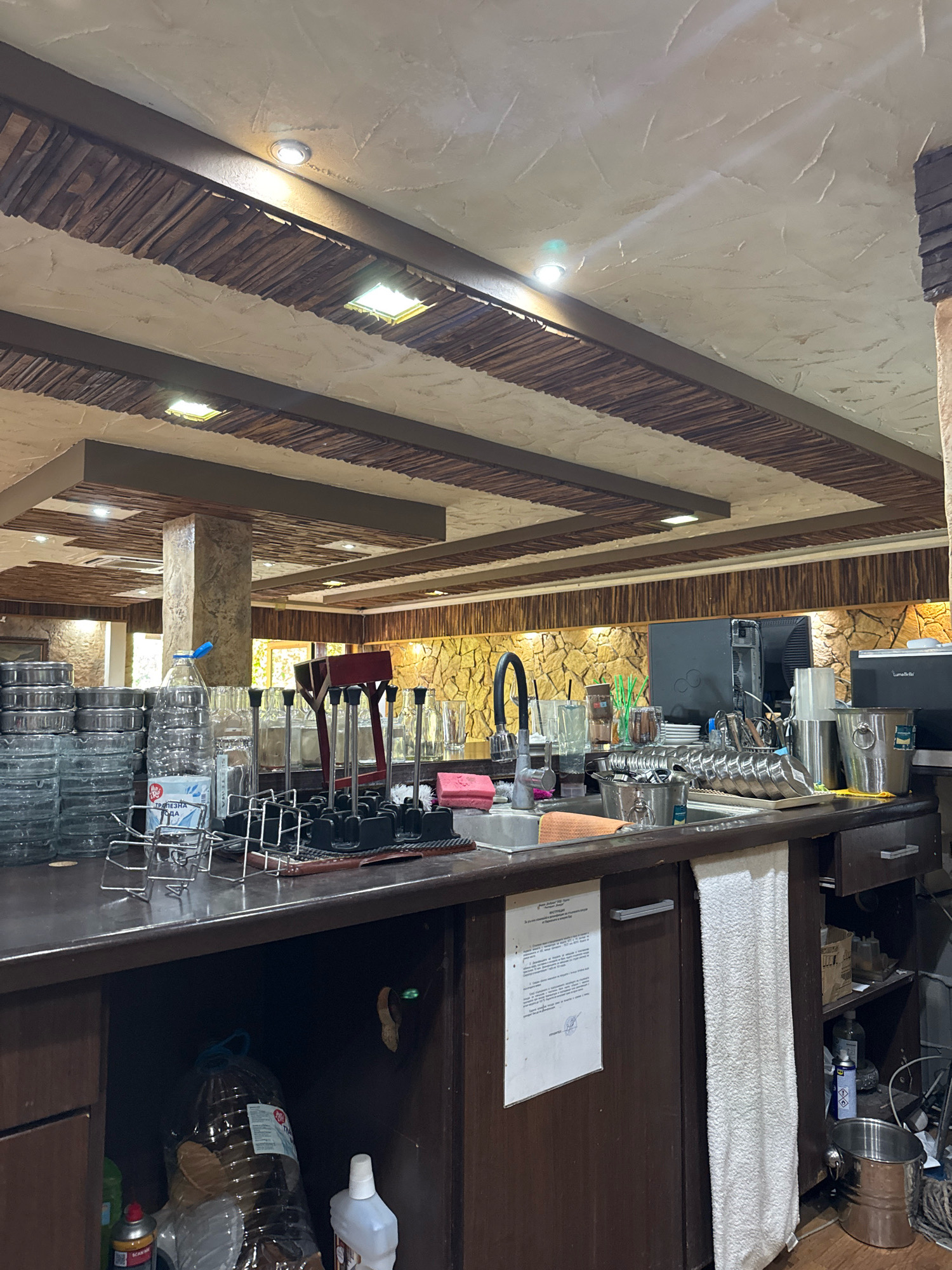 The image depicts the interior of a bar or restaurant in Sredets, Bulgaria. The scene is focused on a bar counter area, with a sink, various glasses, bottles, and bar equipment. The ceiling features wooden beams and recessed lighting. The walls are decorated with stone cladding. In the foreground, stacks of ashtrays are visible on the left, and a water bottle sits on the counter. To the right of the sink, there are glasses, straws, and other bar essentials. Below the counter, cabinets and shelves are partially visible, containing cleaning supplies and large plastic bottles. A white towel hangs from the front of the counter. The overall impression is of a functional service area within a establishment.