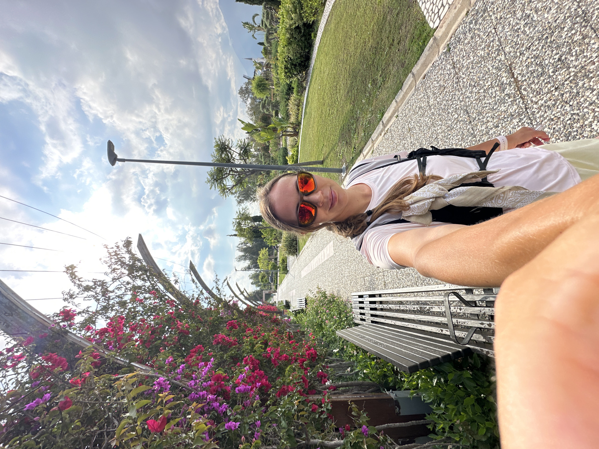 A woman is taking a selfie while sitting on a metal bench in a park in Fethiye, Turkey. The park has a paved walkway and a grassy area with trees and other vegetation. There is also a flower-covered pergola visible. The woman is wearing a white shirt, a backpack, and orange sunglasses. She is smiling at the camera.