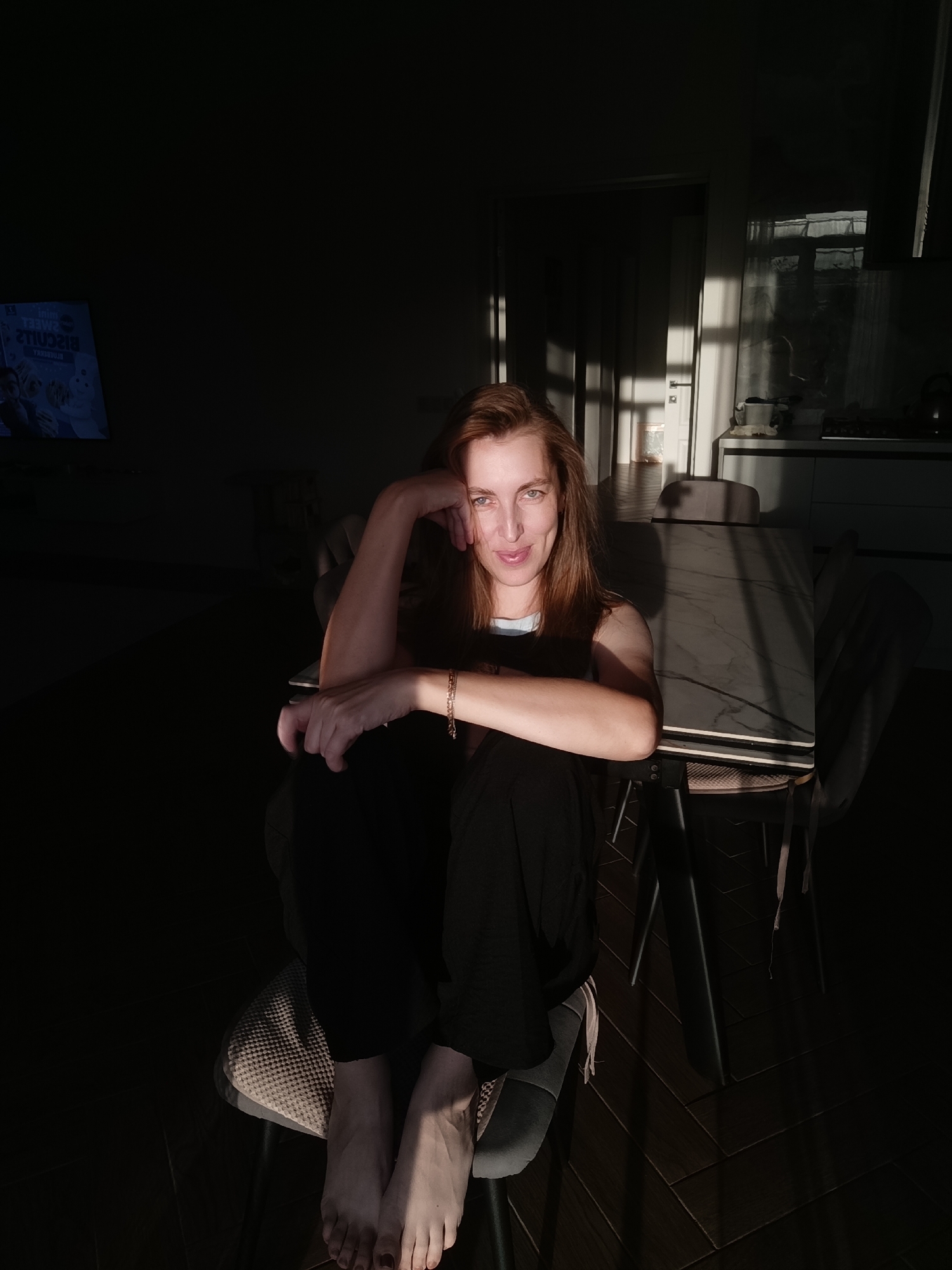 The media file is a vertical, eye-level, full-body shot of a woman sitting on a chair in a room, bathed in the warm, dramatic light of what appears to be late afternoon sun. The light casts long, sharp shadows across the floor and the woman's body, creating a high-contrast, moody atmosphere. The woman is of Caucasian ethnicity with long, light brown hair. She is wearing dark, loose-fitting trousers and a dark top with a light-colored neckline visible. Her legs are crossed, and her feet are bare. She is leaning on her left arm, with her hand resting on her cheek, and her right arm is bent, with her hand resting on her leg. She is looking directly at the camera with a relaxed and slightly enigmatic expression, a hint of a smile on her lips.
The setting appears to be an indoor living space or dining area. To the left of the frame, a television screen is visible, displaying text that is partially obscured by shadow, but some English words like "Sweet" and "Biscuits" are decipherable, suggesting it might be an advertisement or a show. In the background, a table with chairs is visible, and further back, kitchen elements such as cabinets and a countertop are dimly lit. The light filtering in from a window or door creates a strong sense of the time of day, suggesting it is either sunrise or sunset. The overall mood is intimate and contemplative, with the strong interplay of light and shadow emphasizing the woman's presence. The location context is Chaikovsky, Russia.