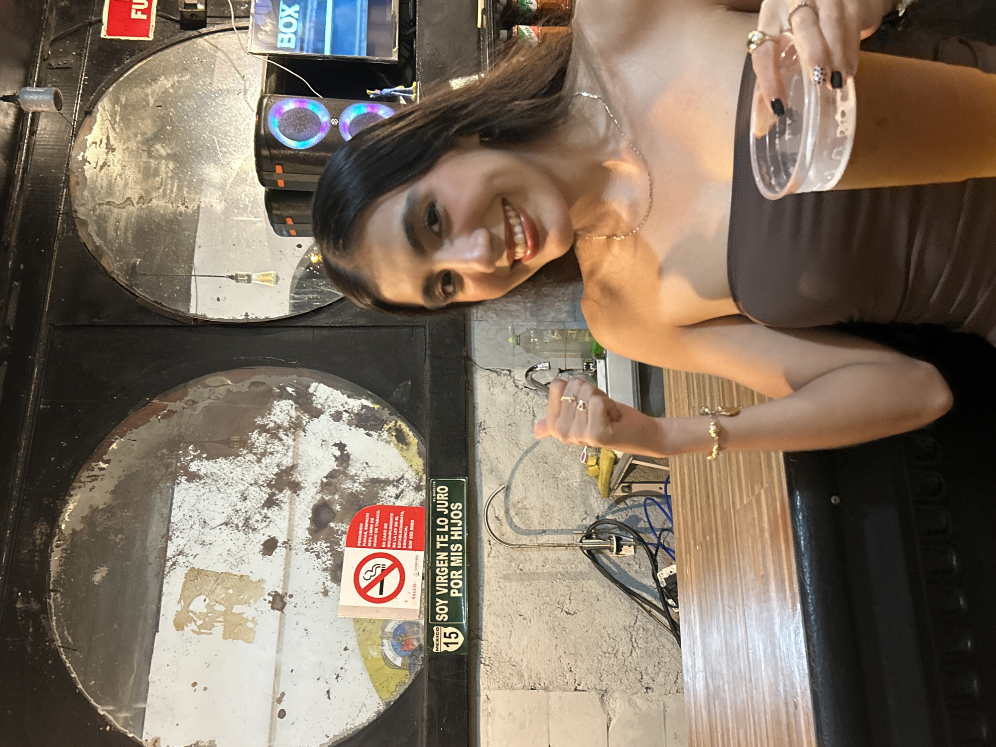 A young woman with long dark hair, wearing a strapless brown top and jewelry, holds a partially filled clear plastic cup containing a yellow liquid (likely beer) and smiles at the camera. She is positioned behind a bar, which is made of light-colored wood.
The background features a wall with exposed concrete and wiring, typical of a casual bar environment. Mounted on the wall, there's a black metal structure with two round, reflective "porthole"-like openings. A round sticker with a no-smoking symbol and the text "PROHIBIDO FUMAR, ESPACIO 100% LIBRE DE HUMO DE TABACO, EN CASO DE INCUMPLIMIENTO DE LA LEY EN EL ESTABLECIMIENTO, DENUNCIA 800 033 5050 SALUD COFEPRIS" is partially attached to one of the openings. A vertical green sign is adjacent to the round metal feature with the text "MEXICO 15 EL QUELITE SOY VIRGEN TE LO JURO POR MIS HIJOS."
Based on the sign "SOY VIRGEN TE LO JURO POR MIS HIJOS," the location is likely in the Quelite area near Mazatlán, Mexico, where such signs are commonly seen in local establishments.