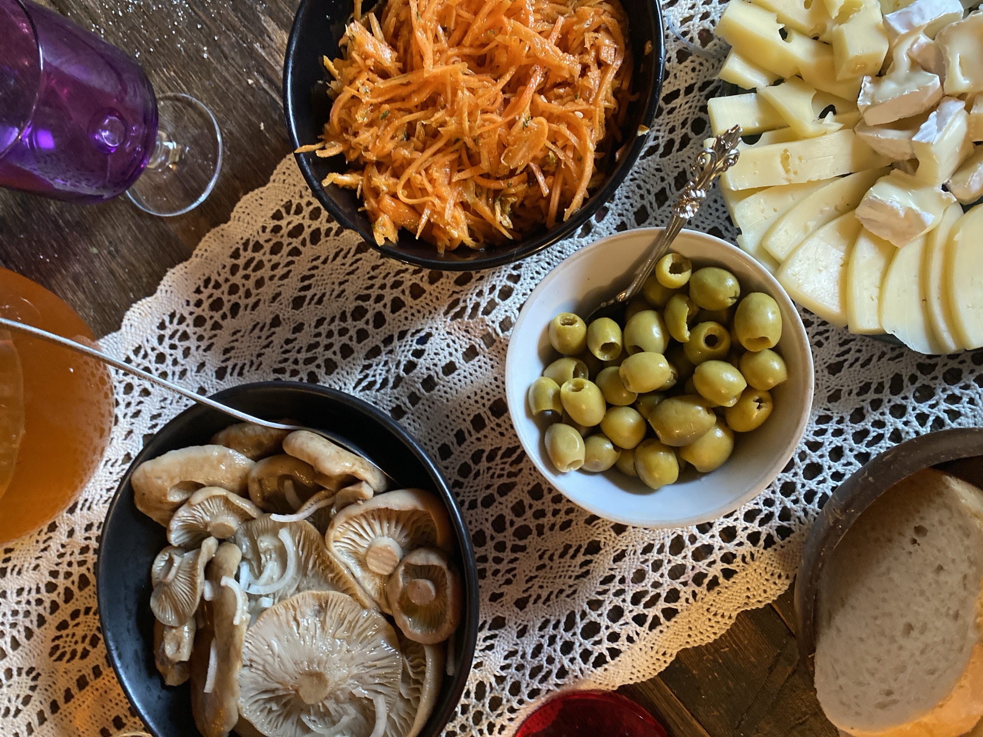 The image shows a close-up, overhead view of a table laden with various food items, set on a dark wooden surface covered by a white, lacy doily. In the top left corner, a purple glass, possibly containing a drink, is tilted. To its right is a black bowl filled with shredded carrots, appearing to be a salad with some herbs. Below the carrot salad is another black bowl containing pickled mushrooms and sliced onions.
To the right of the carrot salad is a white bowl overflowing with green olives, a serving spoon with an ornate handle resting inside. Further to the right, a platter is covered with an assortment of sliced cheeses, including some with holes and creamy white rinds. In the bottom right corner, a slice of bread rests in a dark, round container.
The lighting suggests it might be daytime, possibly indoors, given the controlled lighting and lack of harsh shadows. The overall impression is a rustic and appetizing spread, perhaps a charcuterie board or a selection of appetizers. There is no visible text in the image. The setting appears to be a dining area, possibly a home or a casual restaurant. The focus is entirely on the food and its presentation.