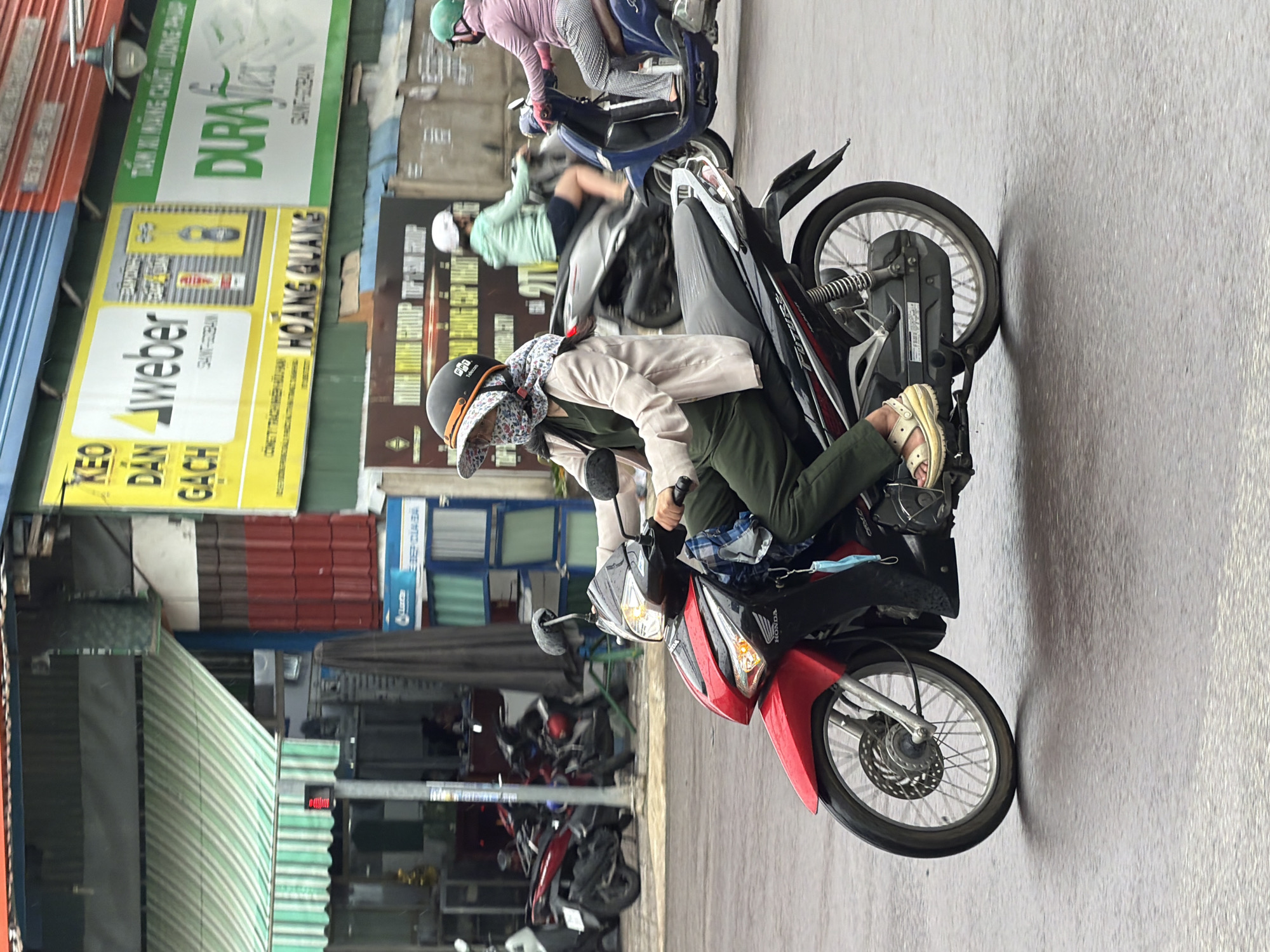 A woman wearing a light-colored jacket, dark green pants, a patterned face covering, and a white and orange helmet is riding a red and black Honda motorcycle. She is wearing light-colored sandals. She is holding onto the handlebars and appears to be driving on a wet, paved road in front of shops and buildings. In the background, other people are riding motorcycles, and there are various signs in Vietnamese. The scene is likely a street in Nha Trang, Vietnam, indicated by the presence of businesses like "DURAFlex" and what appear to be Vietnamese advertisements. The traffic light in the upper left corner of the image is red, suggesting that the woman may be stopped or preparing to stop.