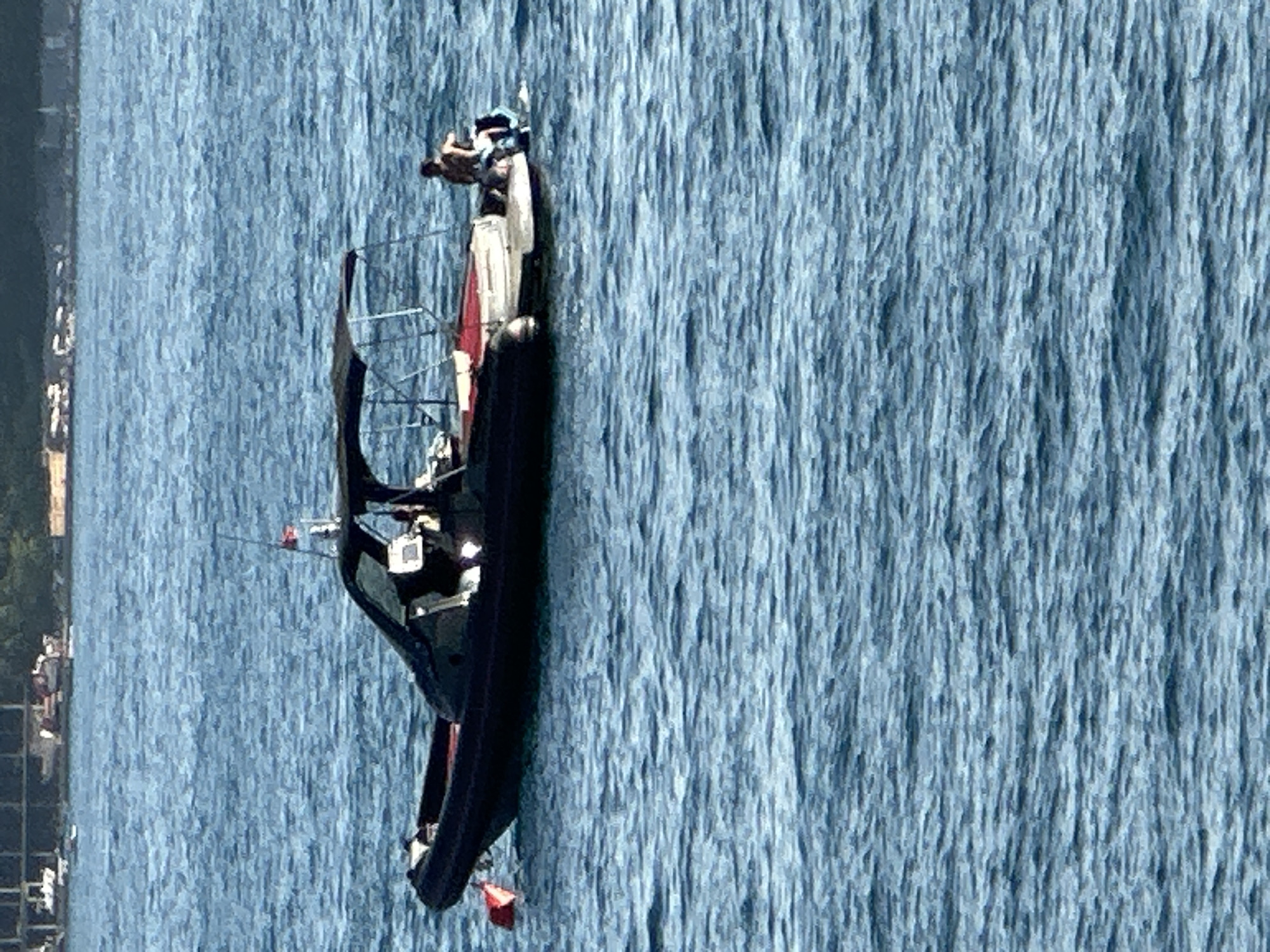 The image shows a black inflatable boat with a white and red cabin floating on a blue, choppy body of water. The boat is oriented diagonally, with the bow pointing towards the bottom left and the stern towards the top right. There are at least two individuals visible on the boat, one in the cabin and another standing near the front.
The scene appears to be outdoors, likely during the daytime, given the bright lighting and reflections on the water. The water's texture suggests it might be a river, lake, or the sea. In the background, on the left side of the image, there are some green trees and buildings, possibly indicating a shoreline or a town. The water has a textured, almost ruffled appearance.
The boat itself has a dark hull, a white superstructure with red accents, and a metal frame or railing. A small red flag is attached to a pole at the stern. The people on board are not clearly discernible, but one appears to be wearing a hat. The overall impression is one of activity on the water, possibly a tour or transport. No visible text is present in the image. The location context provided (Budva, Montenegro) suggests this could be a scene on the Adriatic Sea.