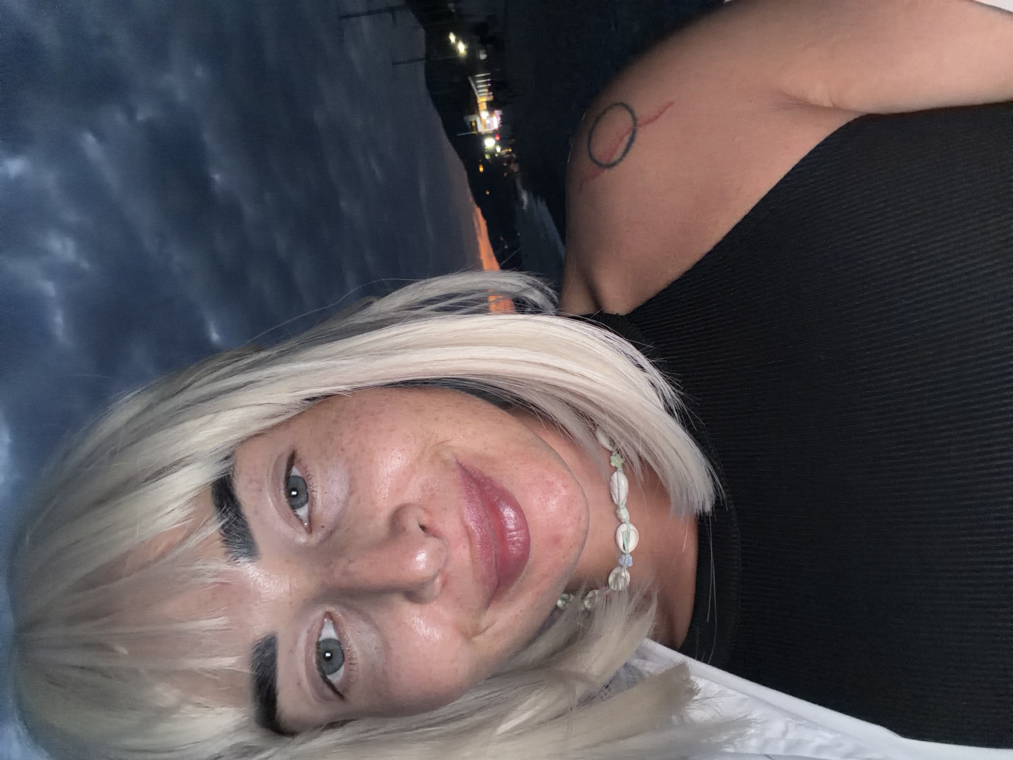 A self-portrait shows a woman with blonde hair and fair skin, wearing a black top and a white shirt. She has a necklace made of shells and small stones. On her left shoulder, a tattoo is visible. The background suggests an outdoor setting, possibly a beach at dusk, with a body of water and distant lights. The sky is dark and cloudy. It is not possible to determine the specific location within the city of Sochi, Russia, from this image alone.