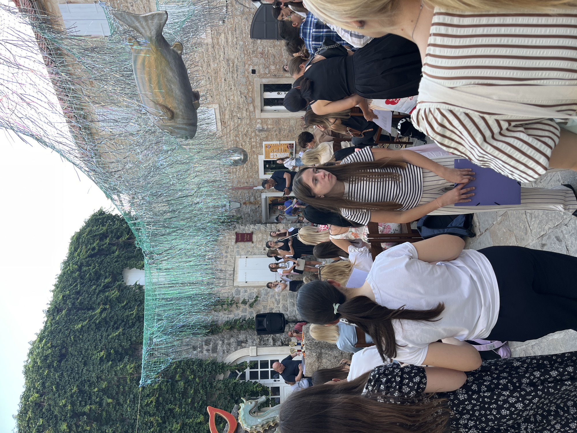 The image shows an outdoor event taking place in what appears to be a courtyard. Several people are seated in rows facing a central area with a speaker and some trophies. Many are looking towards the speaker. A woman in a striped shirt is holding a purple folder.
Above the scene hangs a large decorative installation featuring a sculpture of a fish and netting. The buildings surrounding the courtyard are covered in vines. The event is taking place in Budva, Montenegro.