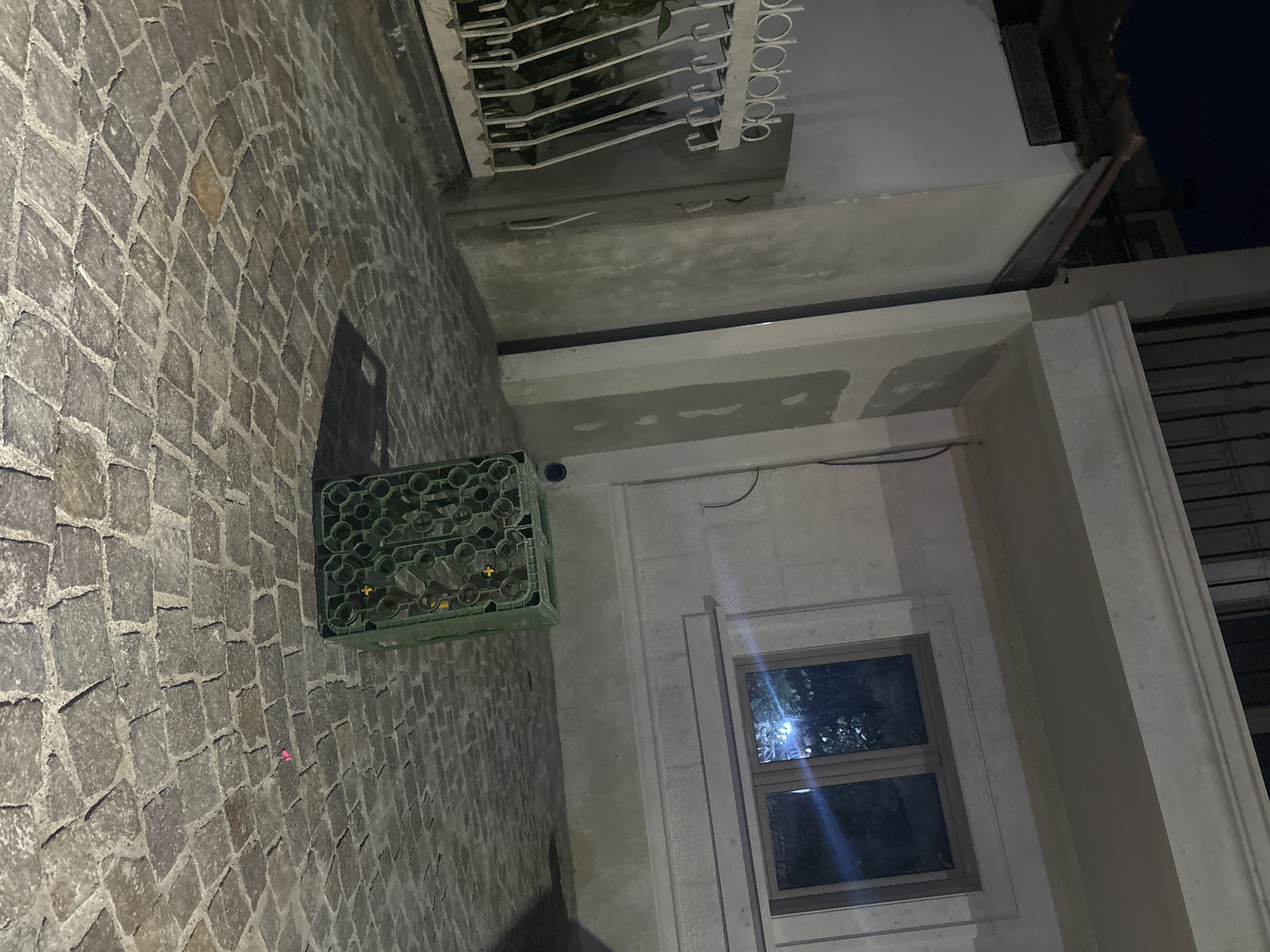 In the city of Budva, Montenegro, an outdoor scene unfolds at night. The foreground is dominated by a cobblestone ground, laid in a circular pattern. In the center of the image, a green plastic crate, likely for bottles, rests on the cobblestones. The crate appears to be filled with items, possibly bottles, indicated by circular indentations. To the right of the crate, a small, bright pink object, possibly a piece of litter or a small toy, lies on the ground.
Behind the crate, a building with modern architecture is visible. The building has a cream-colored facade and a prominent white horizontal molding. A window on the building's facade reflects ambient light, showing hints of foliage and artificial lighting within. A vertical concrete pillar supports an upper structure. To the left of the pillar, a white metal fence with a decorative pattern encloses a small garden area with plants. The overall lighting suggests nighttime, with artificial lights illuminating the scene.